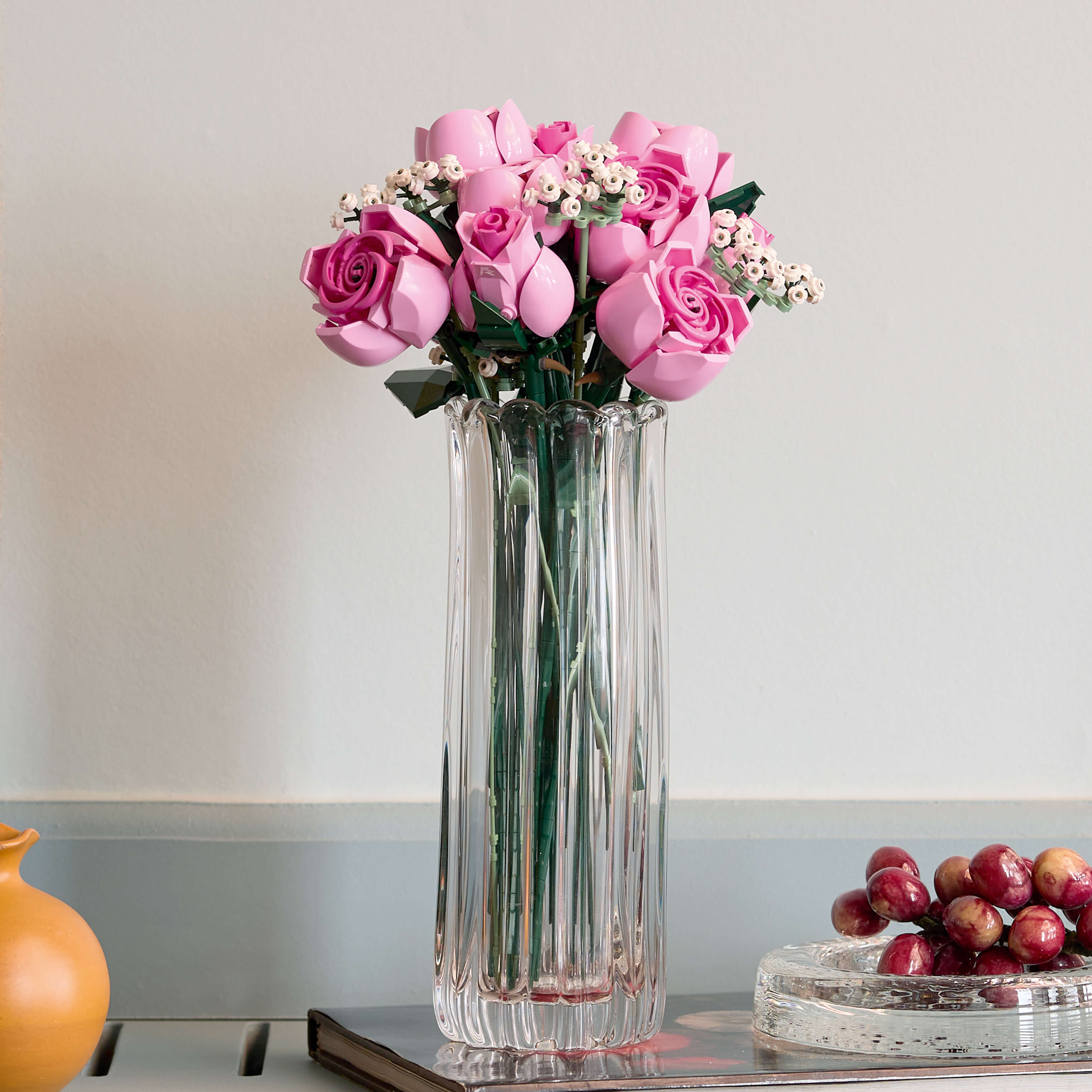 Bukiety różowych róż w szklanym wazonie stoją obok miski z czerwonymi winogronami.