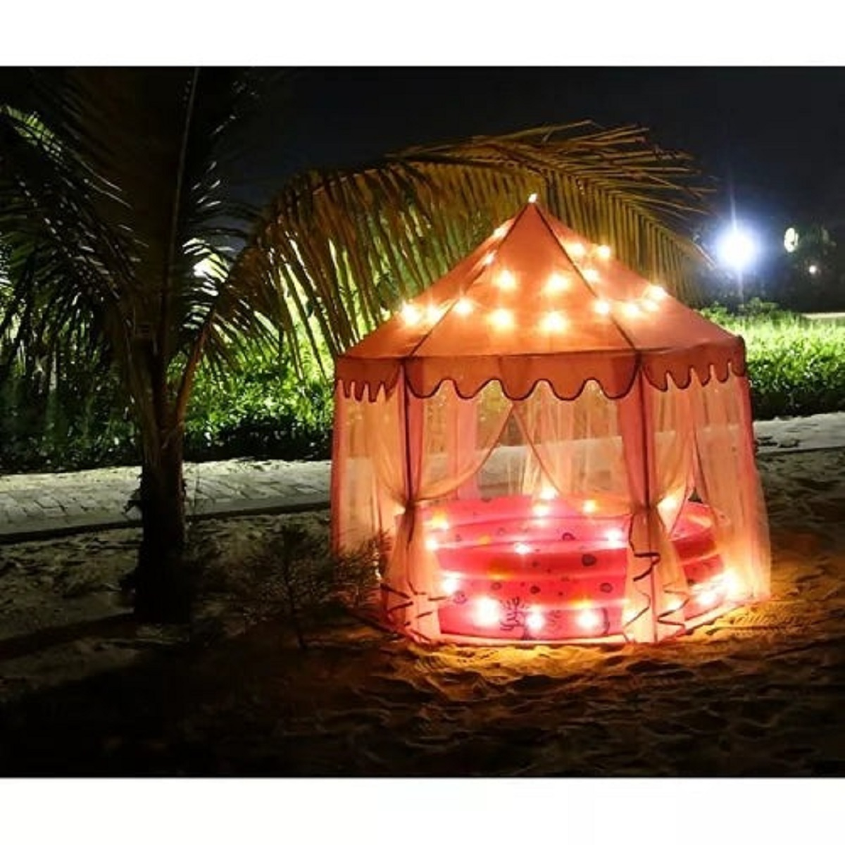 Una tienda rosa iluminada se asienta en la arena junto a una palmera por la noche.