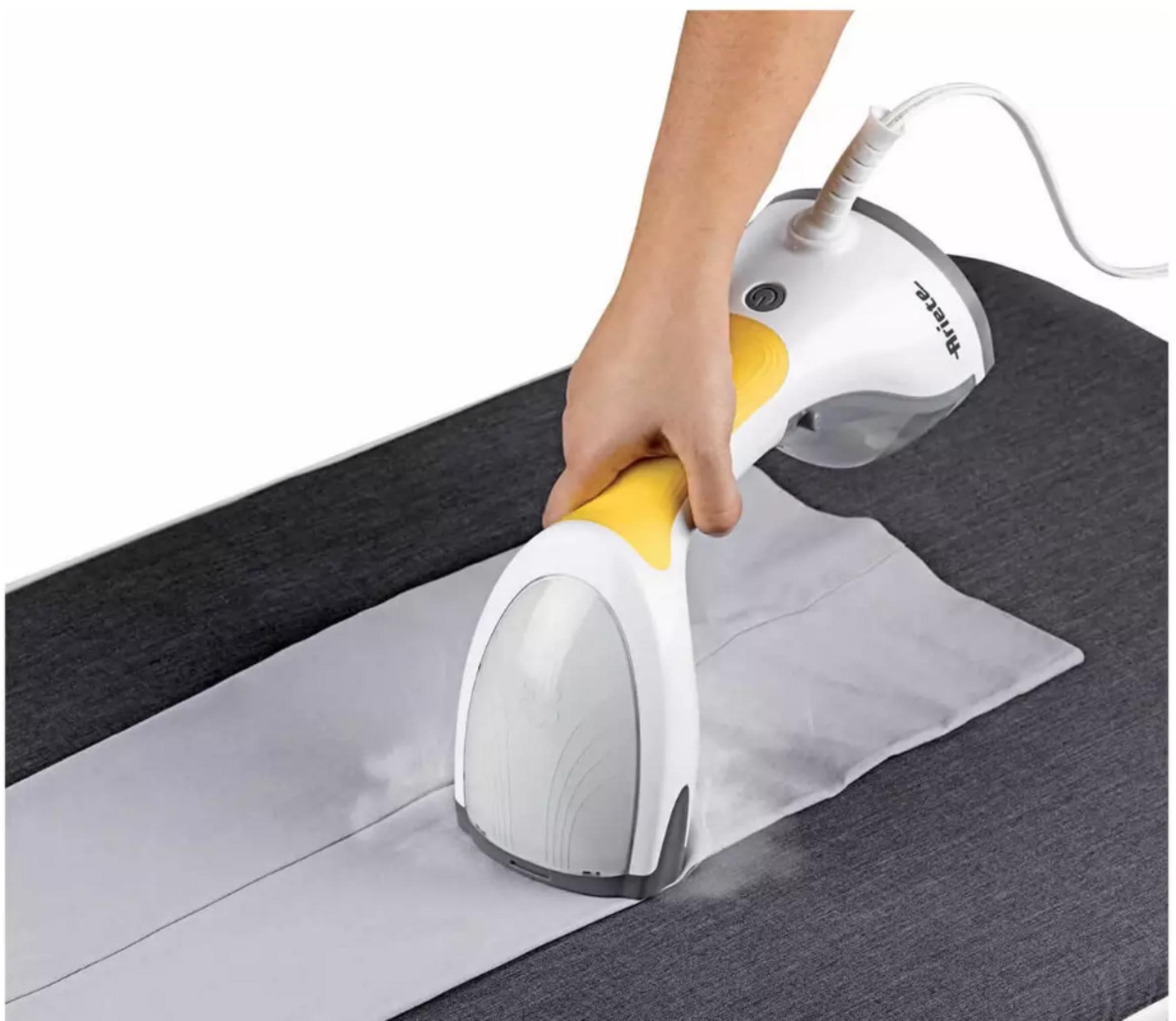 Ręka trzyma biało-żółty parownicę do ubrań na szarej desce do prasowania.