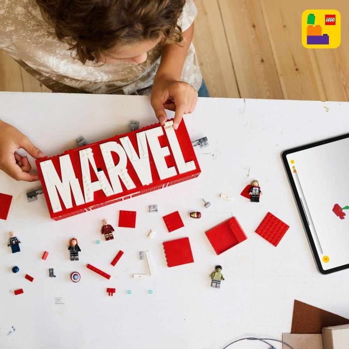 Dziecko buduje logo Marvel z czerwonych i białych klocków LEGO. Wokół leży kilka minifigurek.