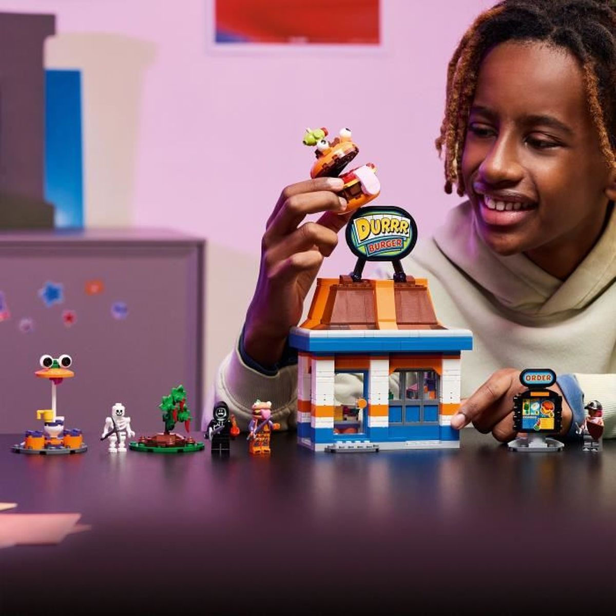 Bir çocuk, figürler ve kurbağa oyuncağı ile Lego burger dükkanı seti yapıyor.