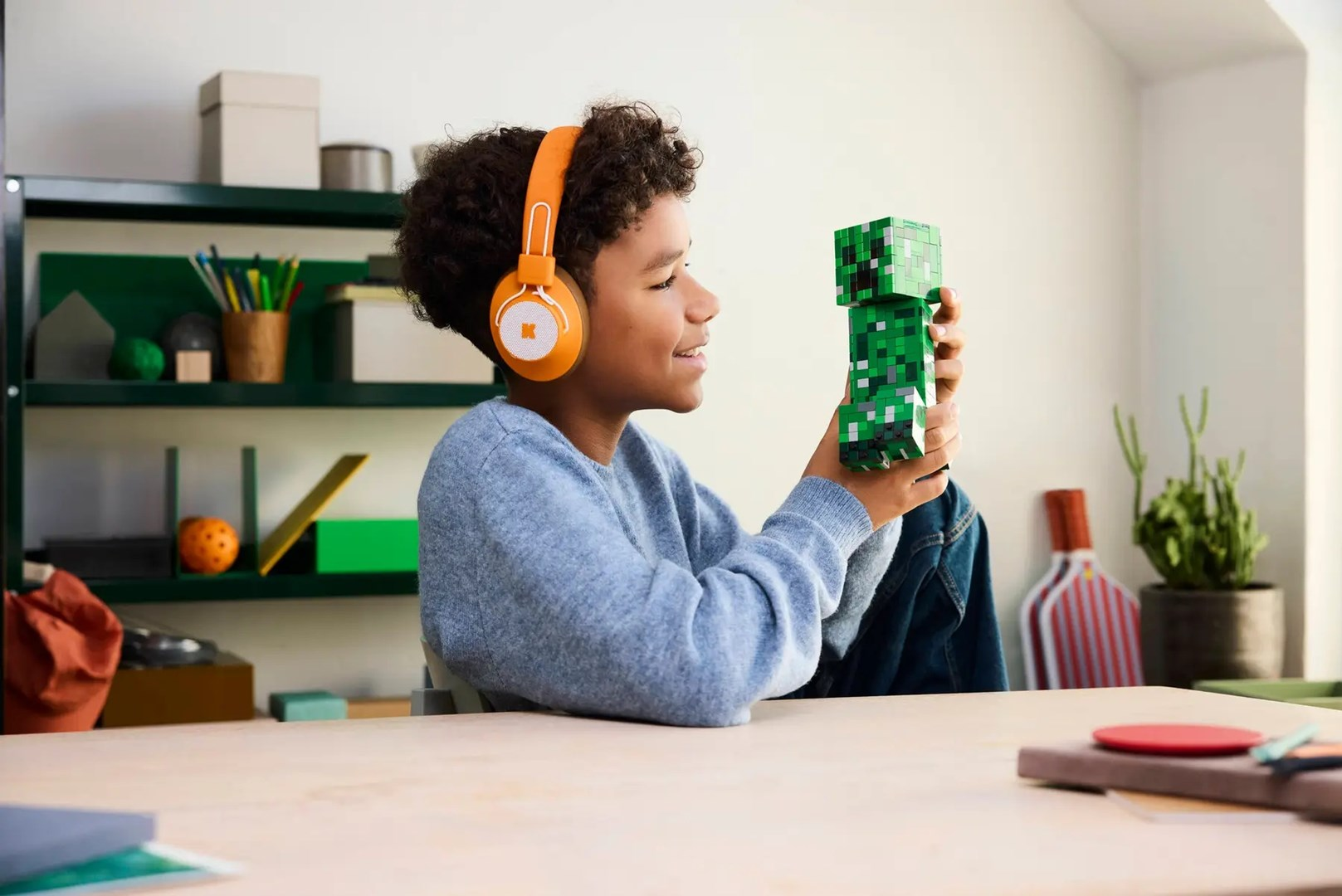 Chłopiec w pomarańczowych słuchawkach trzyma zieloną figurkę Minecraft.