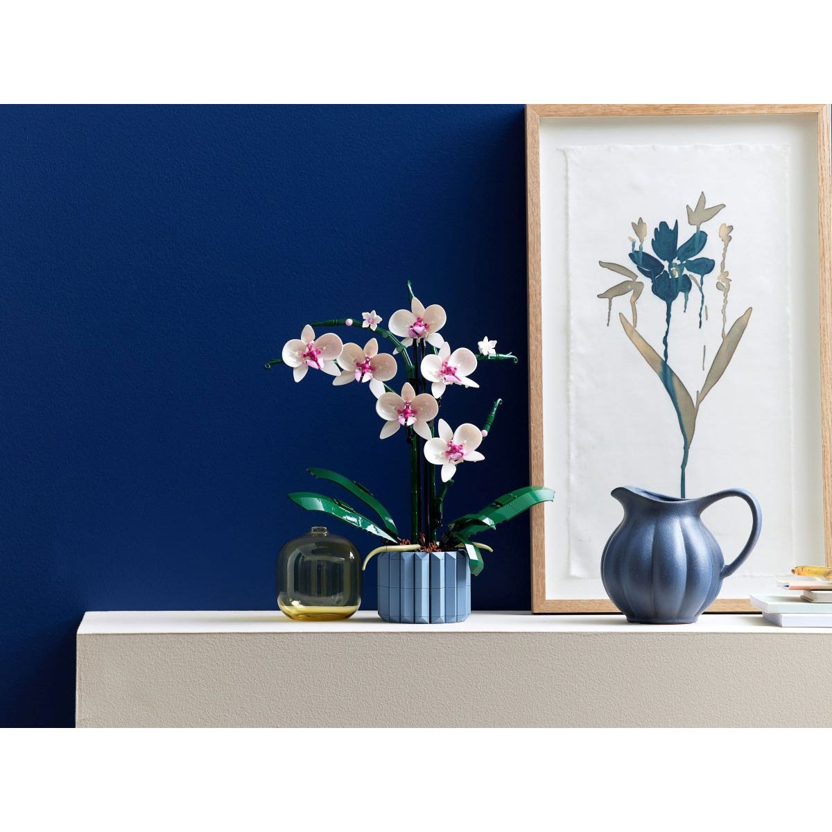 Biała orchidea w niebieskiej doniczce obok dzbanka i dzieła sztuki.
