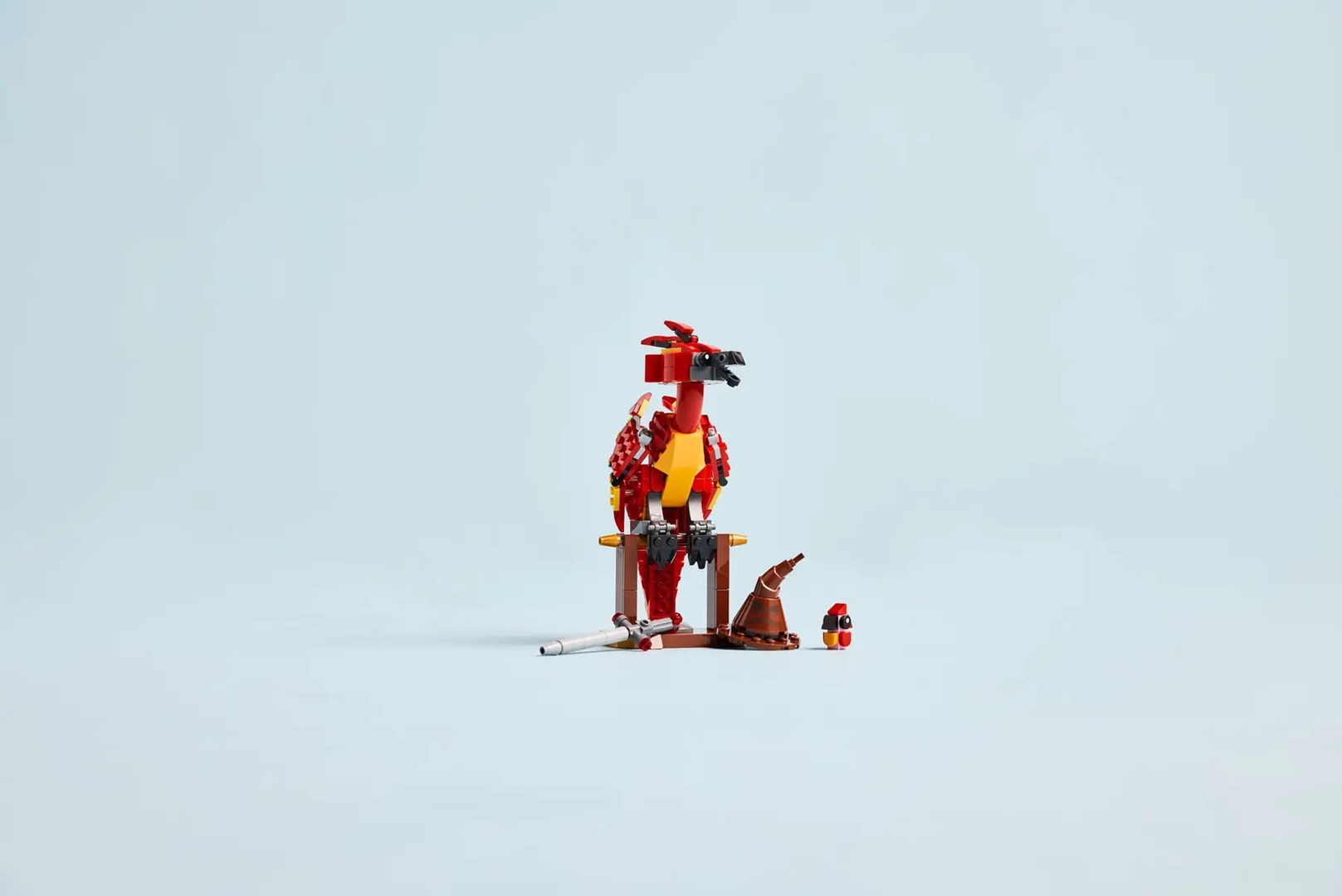 Czerwony ptak Lego siedzi na drewnianym patyku z kapeluszem i figurką obok.