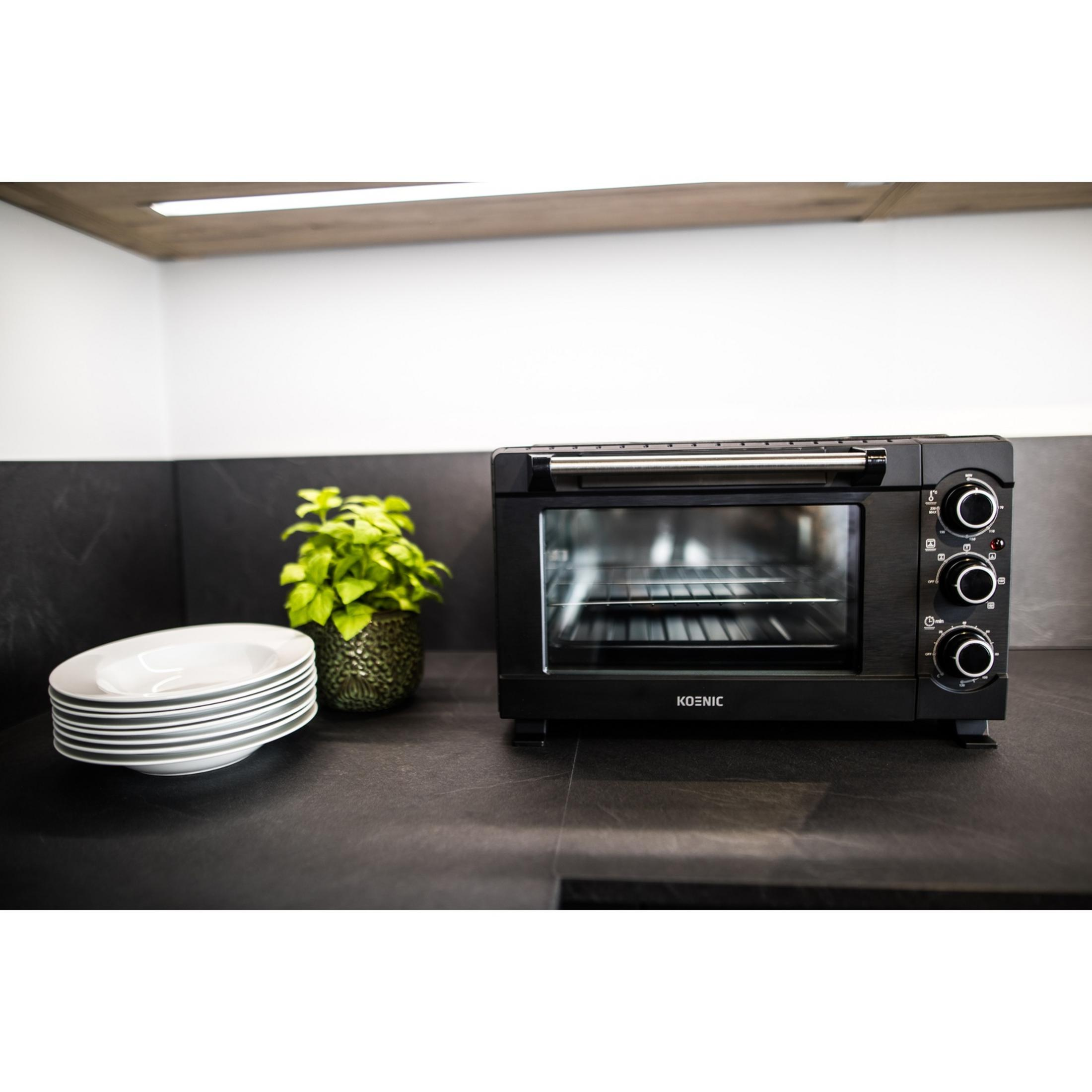 Horno tostador negro sobre encimera oscura con platos y planta.