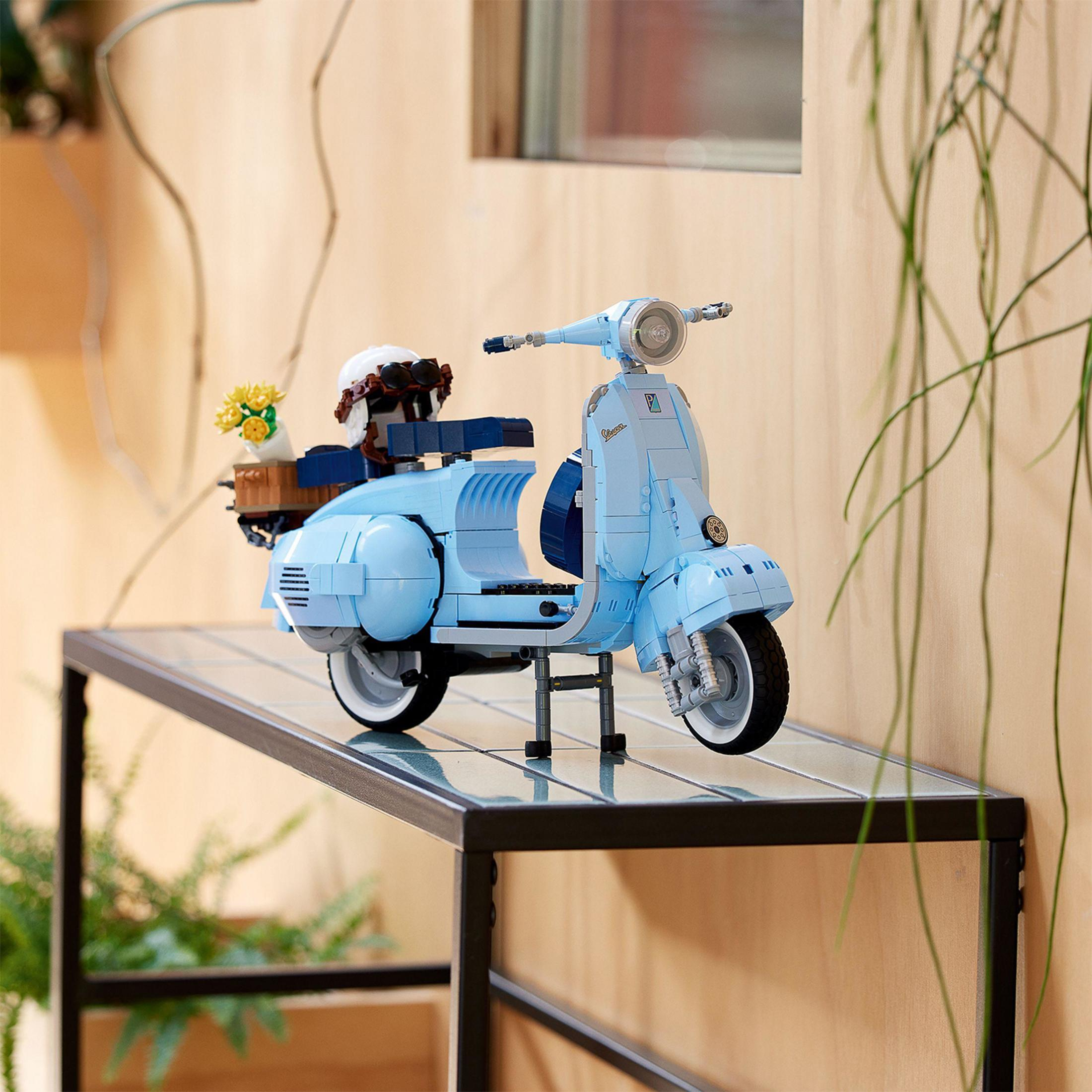 Jasnoniebieski skuter Vespa Lego stoi na półce. Kwiaty w koszu.
