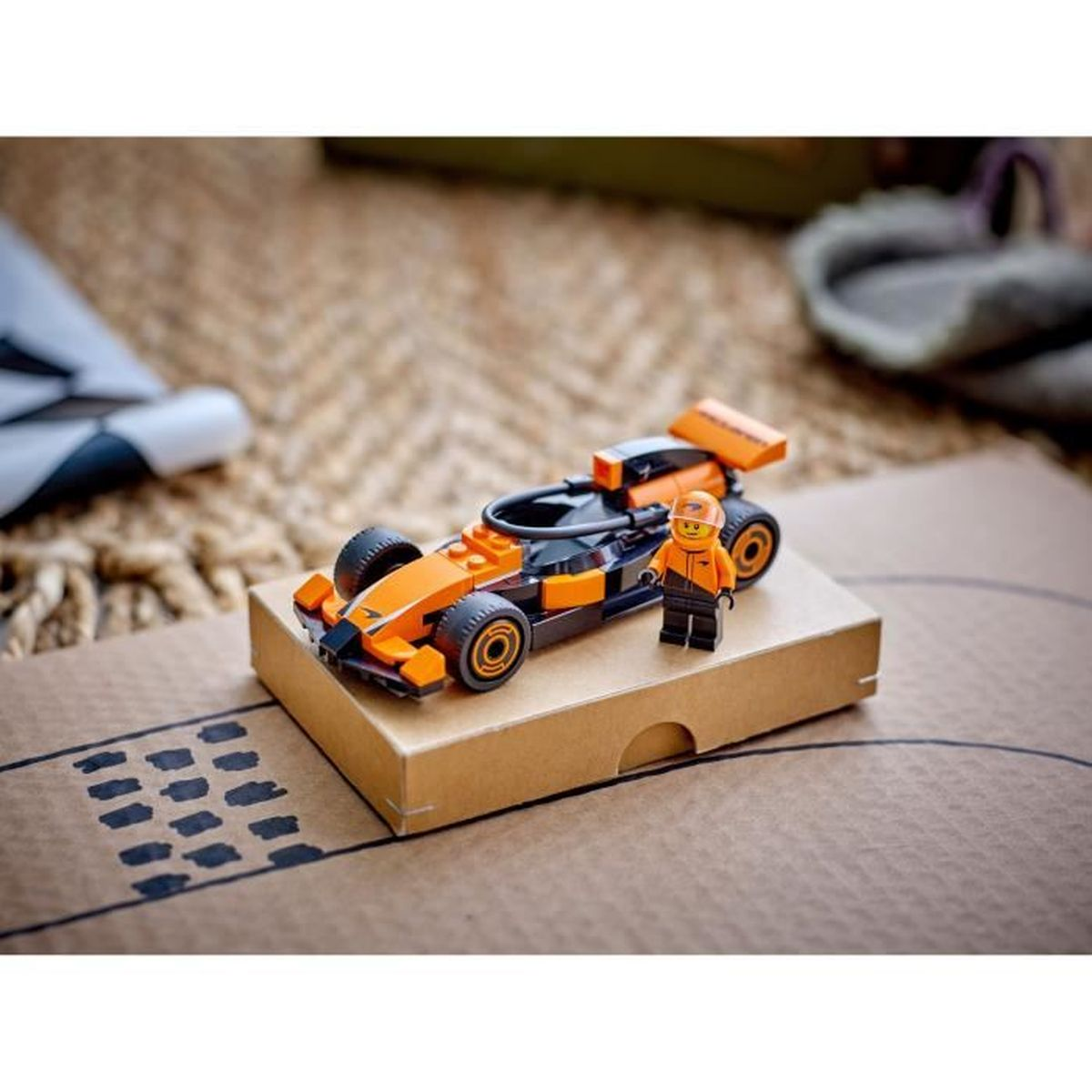 Een oranje Lego raceauto met een coureur naast een kartonnen doos.