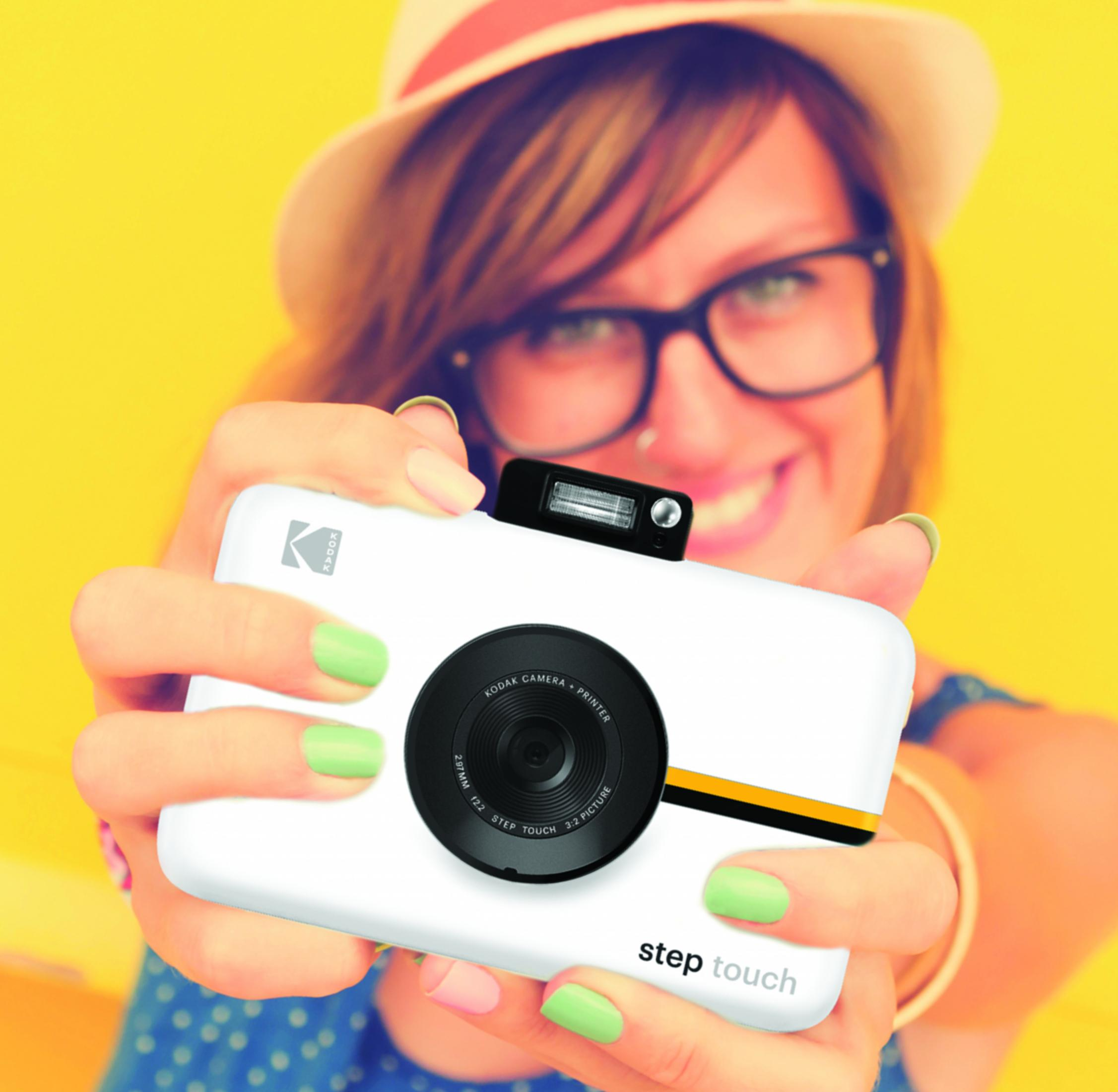 Frau mit Brille und Hut hält Kodak-Kamera mit hellem Hintergrund.