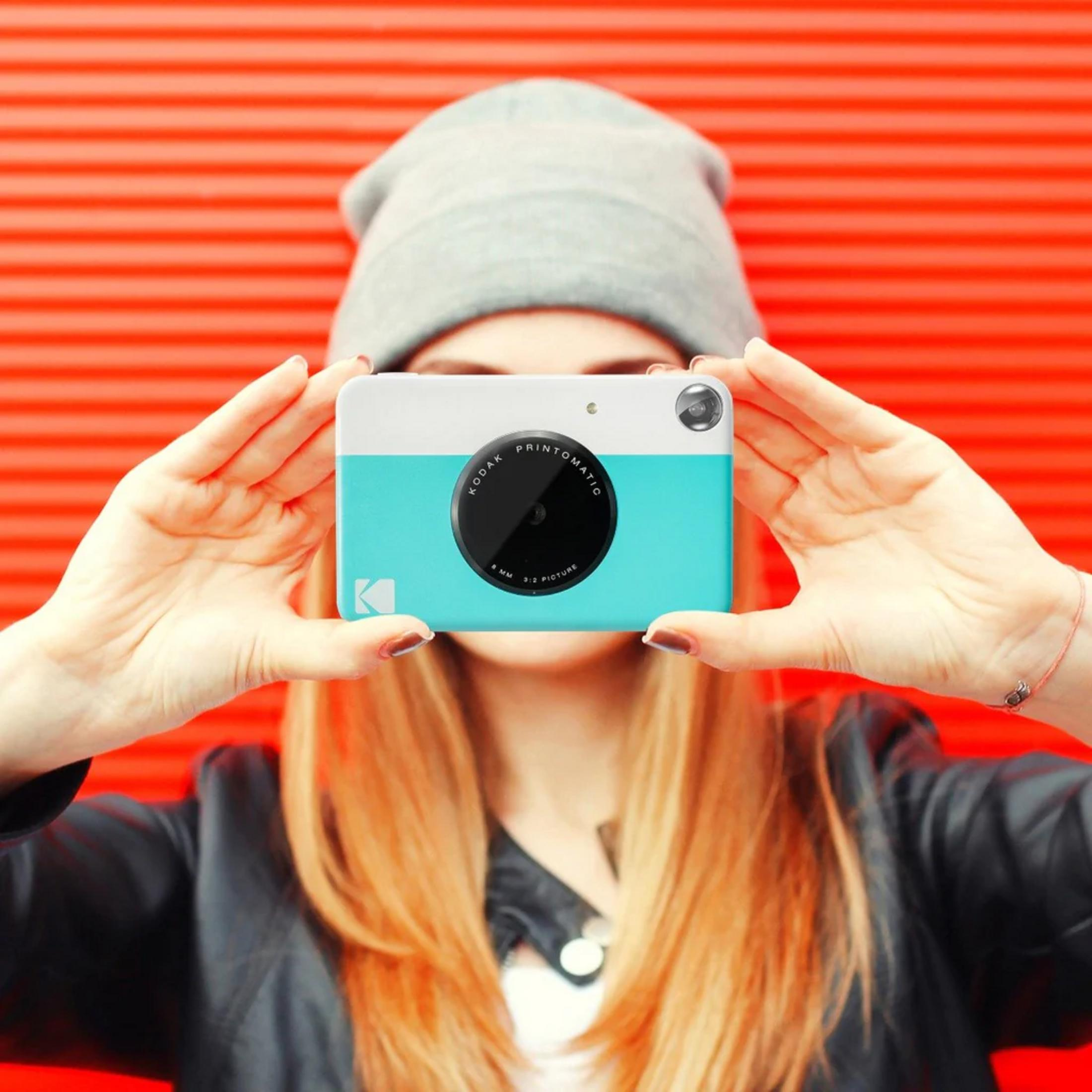 Personne tenant un appareil photo Kodak blanc et turquoise. Fond rouge.