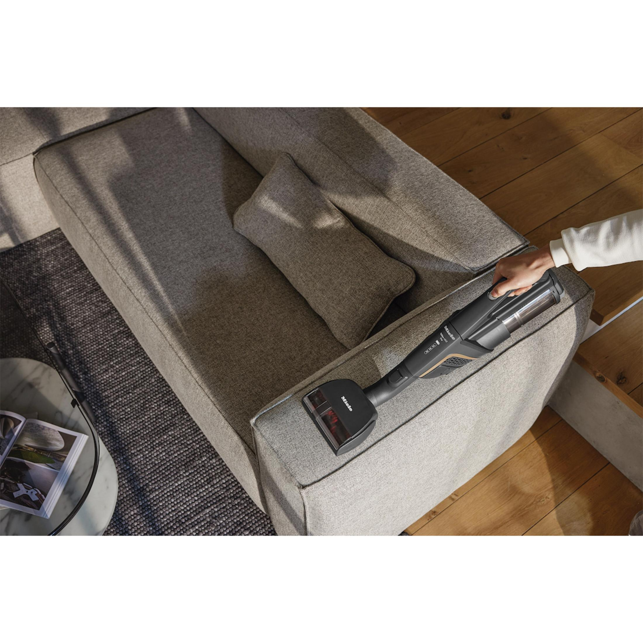 Ręka używa czarnego odkurzacza do czyszczenia szarej sofy.