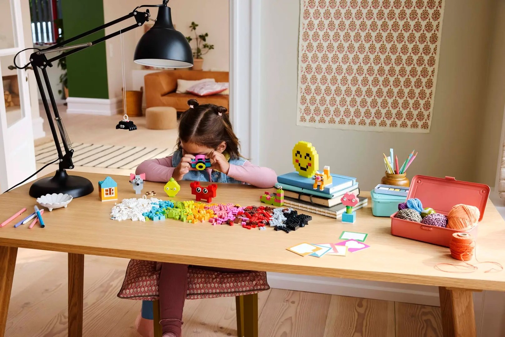 Dziecko bawi się kolorowymi klockami na drewnianym stole z lampą i materiałami plastycznymi.
