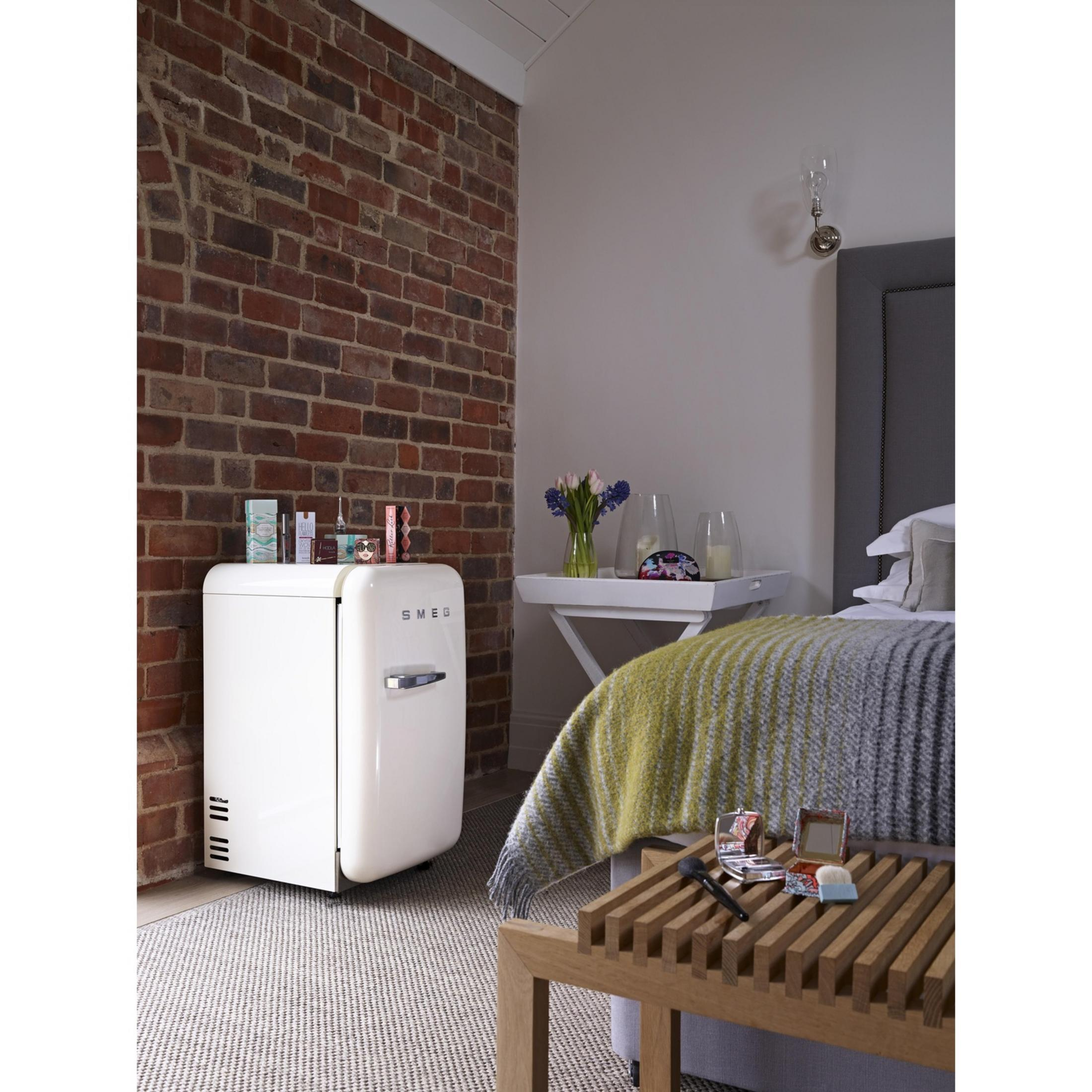 Weiße Smeg-Minikühlschrank neben Backsteinwand mit Bett und Blumen.