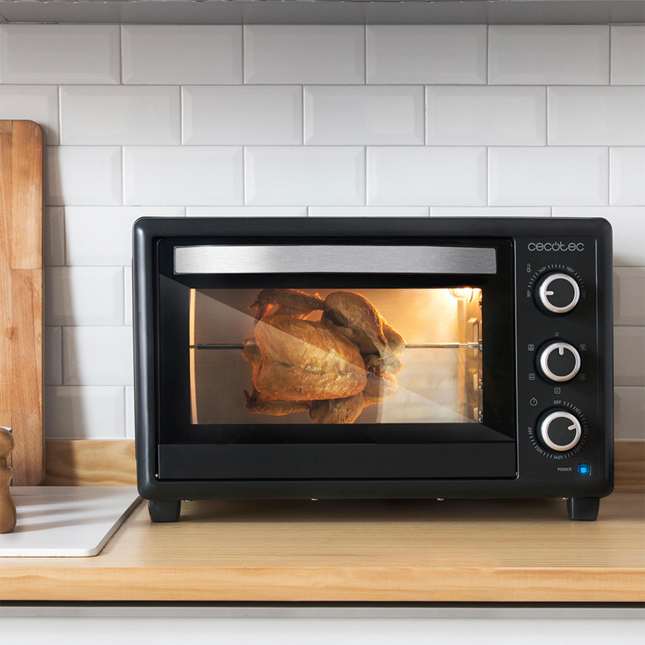 Un pollo intero arrostisce su uno spiedo all'interno di un piccolo forno nero su un bancone di legno.