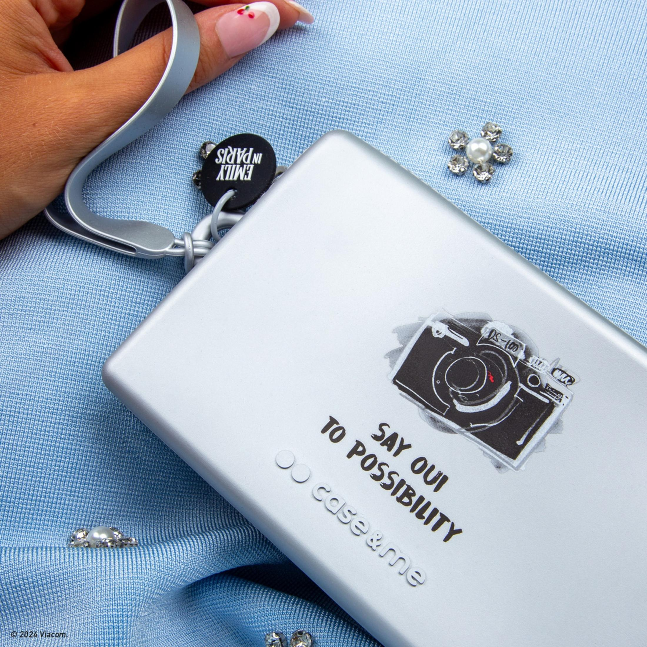 Funda plateada con gráfico de cámara. Texto: 'SAY OUI TO POSSIBILITY'.