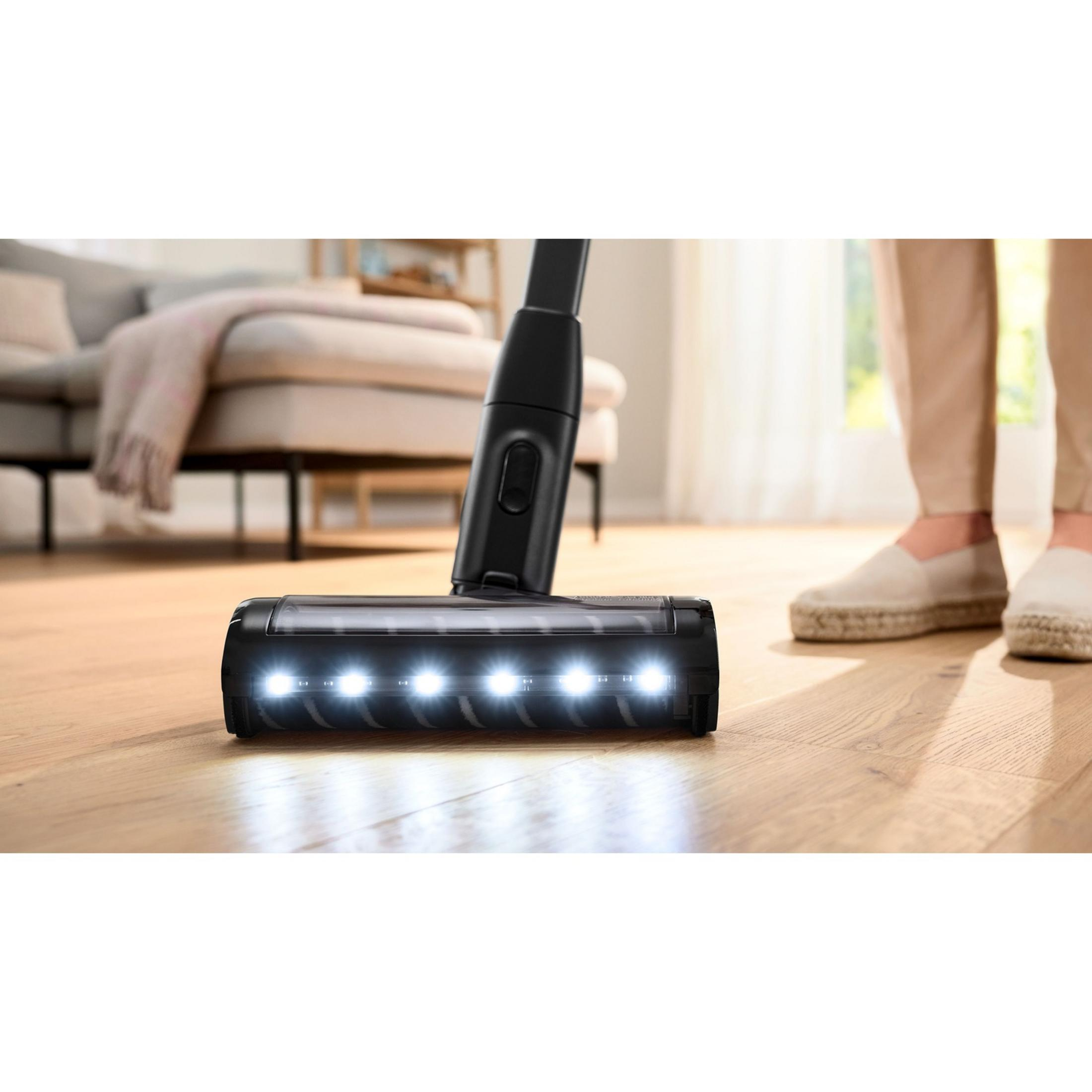 Boquilla de aspiradora con luces sobre suelo de madera, persona al fondo.
