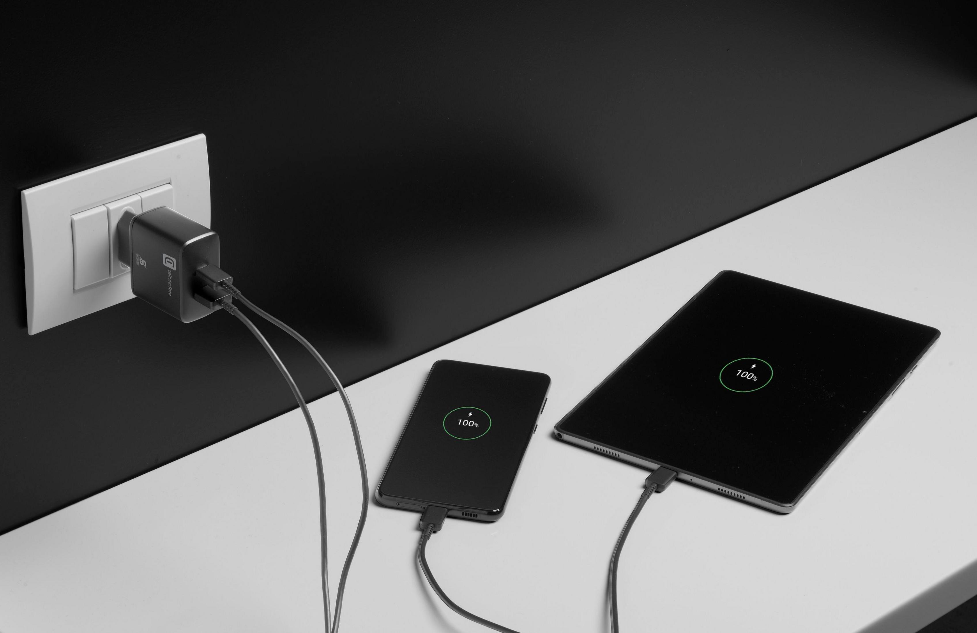Ein schwarzer Ladestecker ist in eine weiße Steckdose gesteckt und lädt ein Smartphone und Tablet.