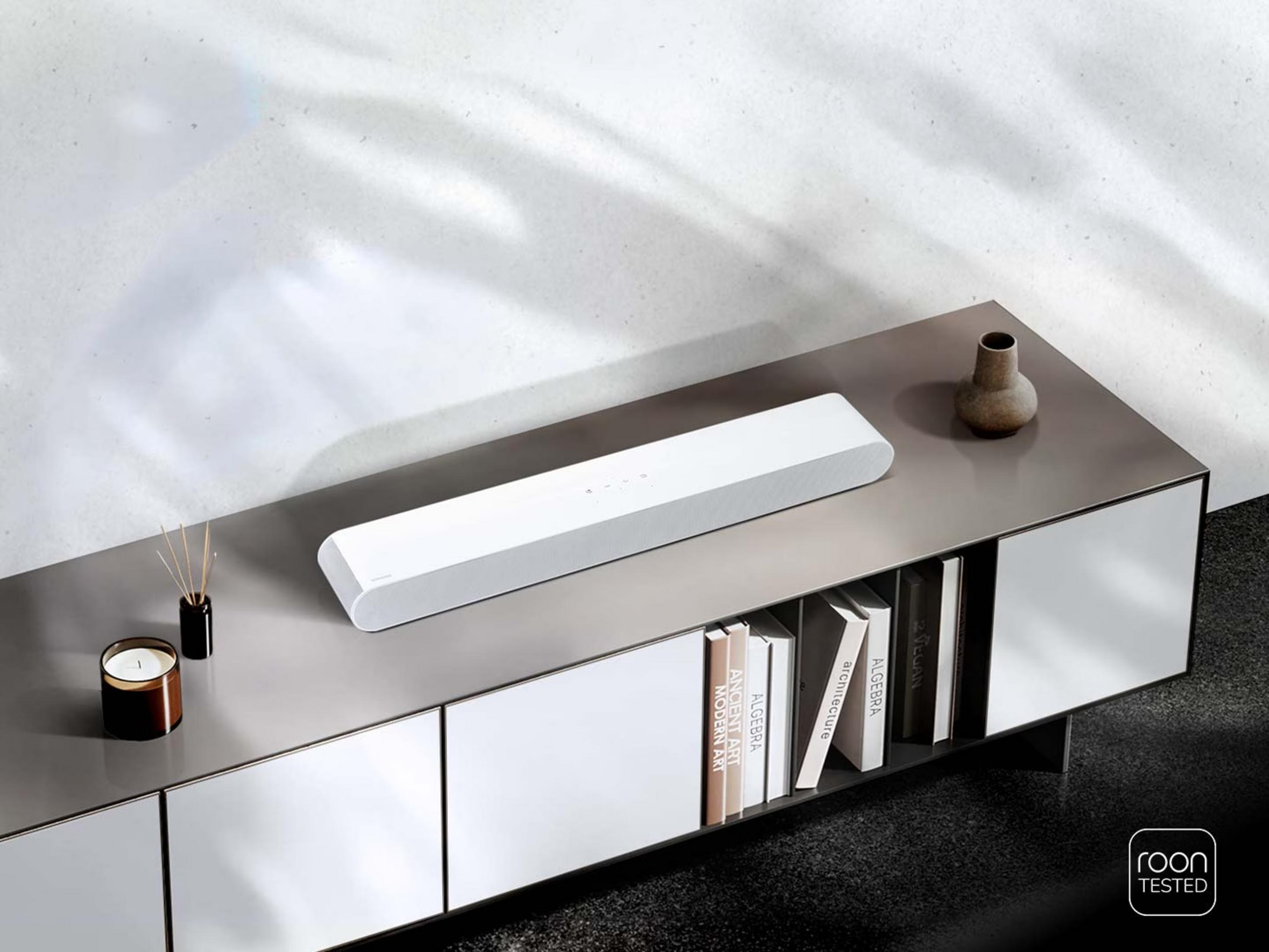 Barre de son blanche sur console grise avec livres et déco. Logo Roon Tested.