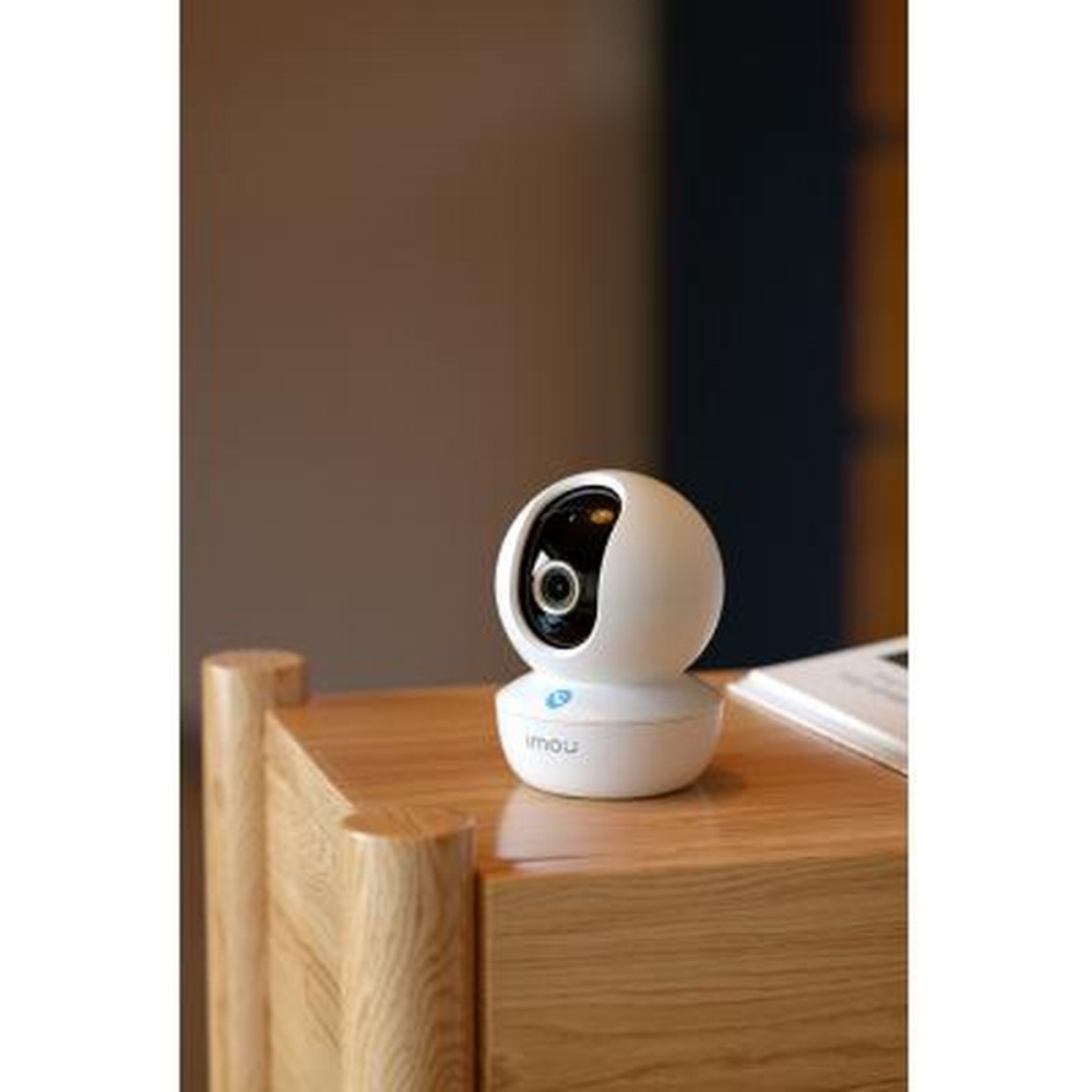 Biała kamera bezpieczeństwa z niebieskim logo stoi na drewnianym stole.