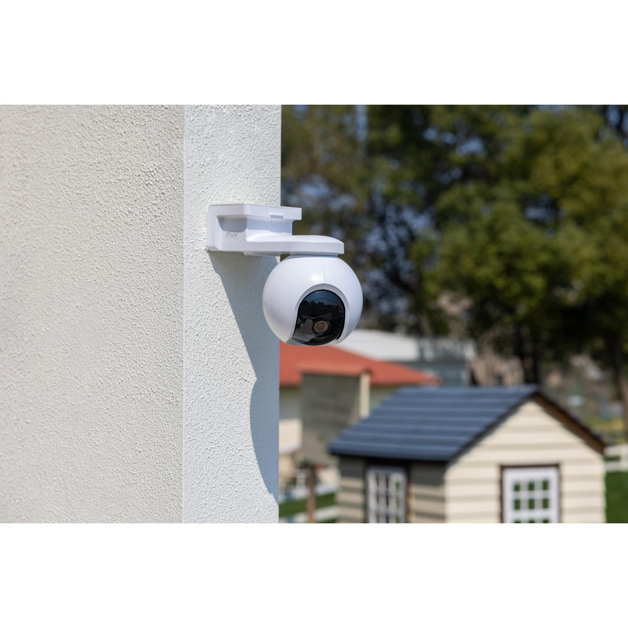 A white security camera is mounted on a wall. A playhouse is in the background.