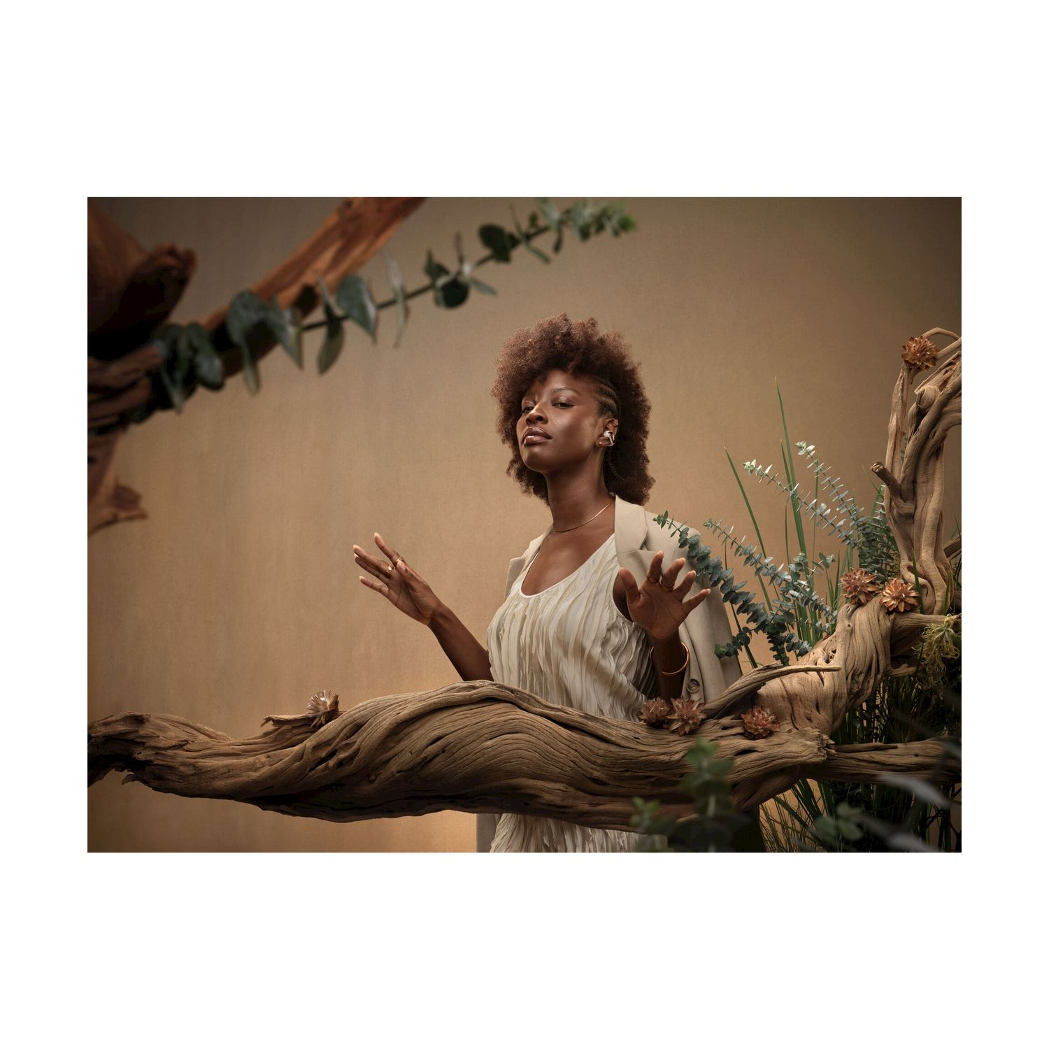 Una donna con capelli afro sta davanti a uno sfondo marrone, circondata da legno e piante.