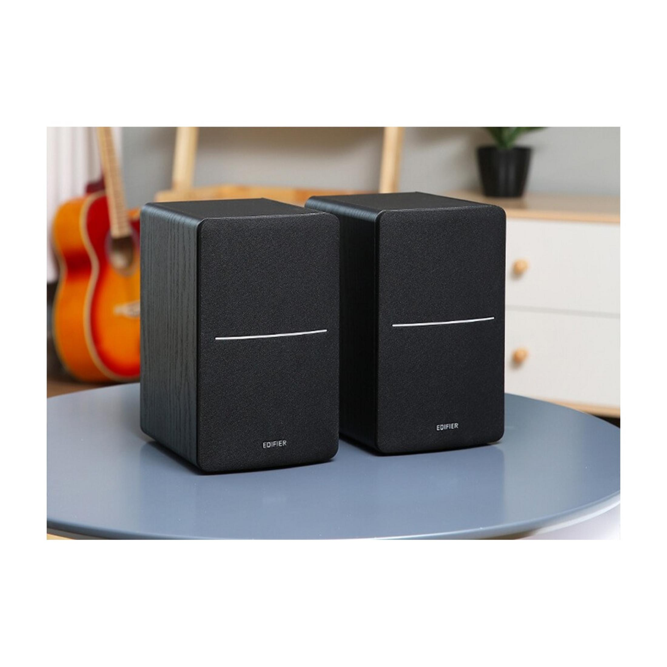 Dwa czarne głośniki na stole, z gitarą i rośliną w tle.