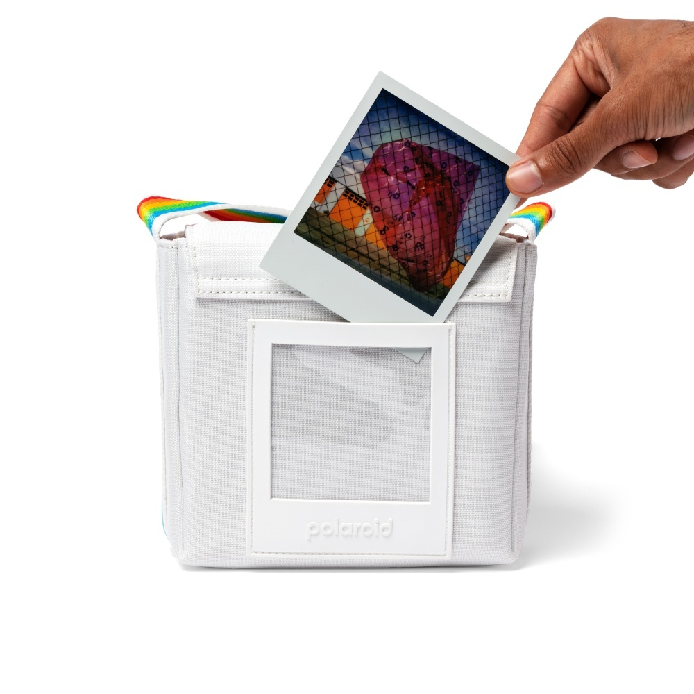 Eine Hand hält ein Polaroidfoto über einer weißen Tasche mit Regenbogen-Trägern.