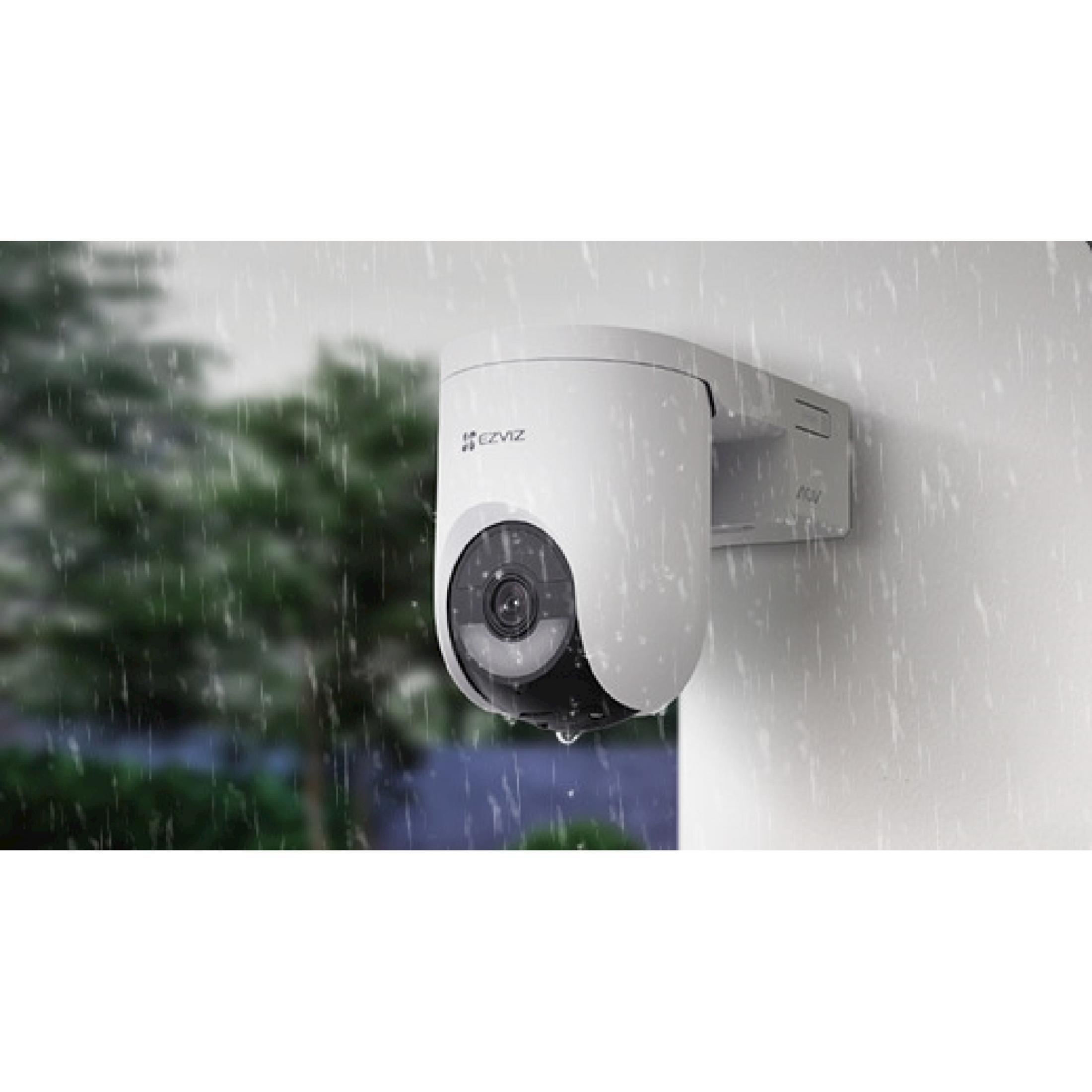 Weiße Überwachungskamera an einer Wand bei Regen, mit Bäumen im Hintergrund.