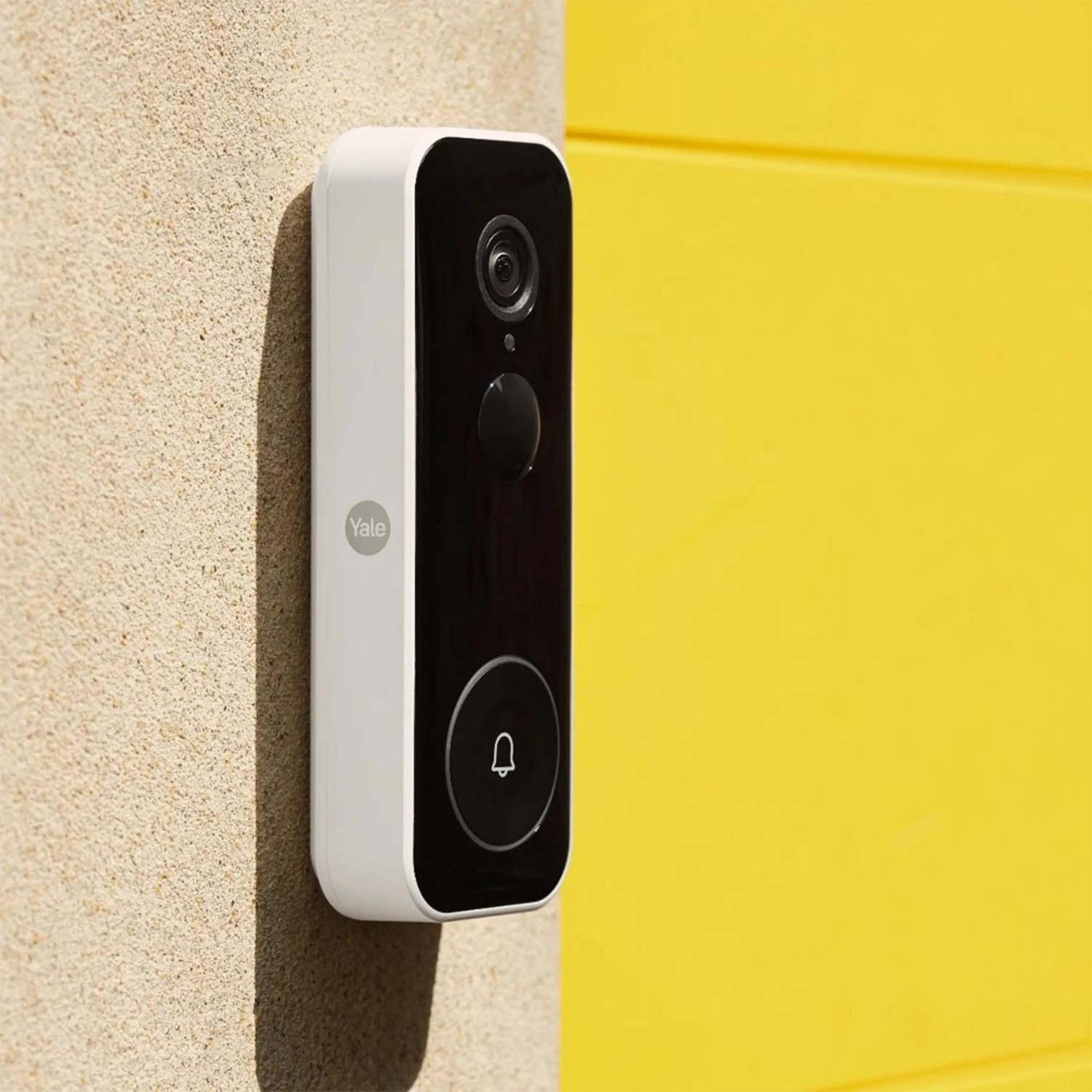 Un timbre Yale blanco con cámara montado en pared texturizada junto a puerta amarilla.