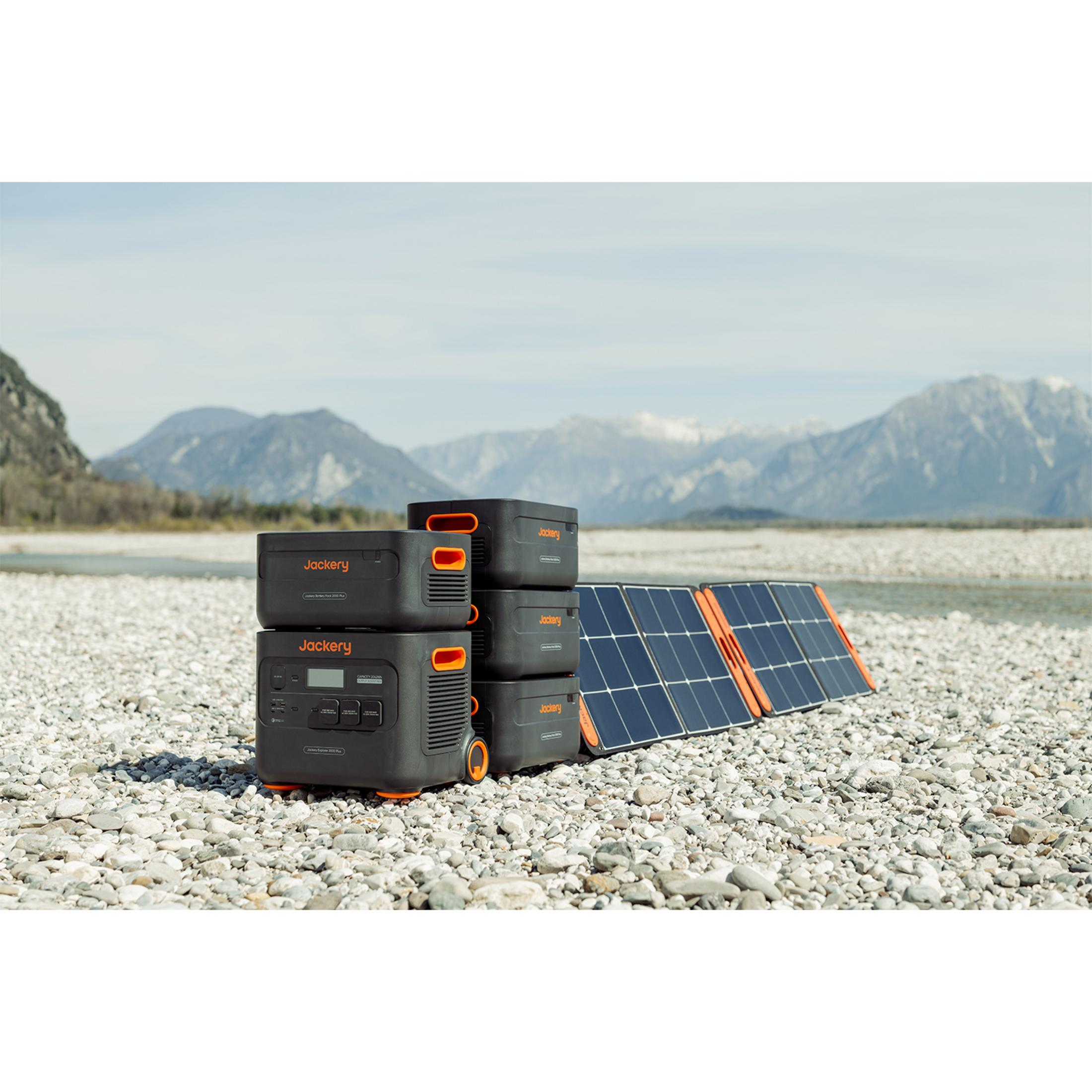 Quattro centrali elettriche Jackery e due pannelli solari su una spiaggia di ciottoli davanti alle montagne.