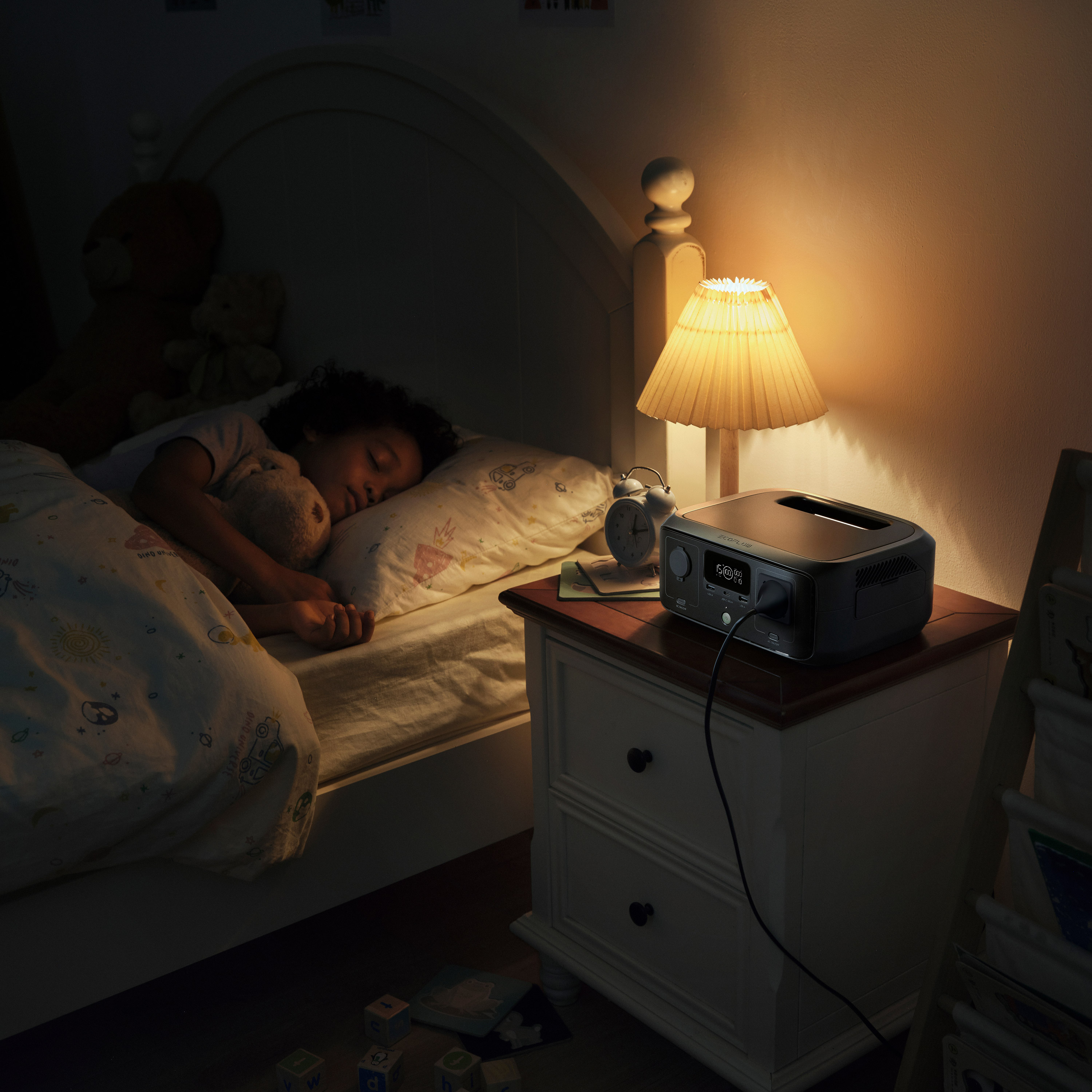 Dziecko śpi w łóżku z pluszowymi misiami. Szafka nocna z lampą i stacją zasilania.