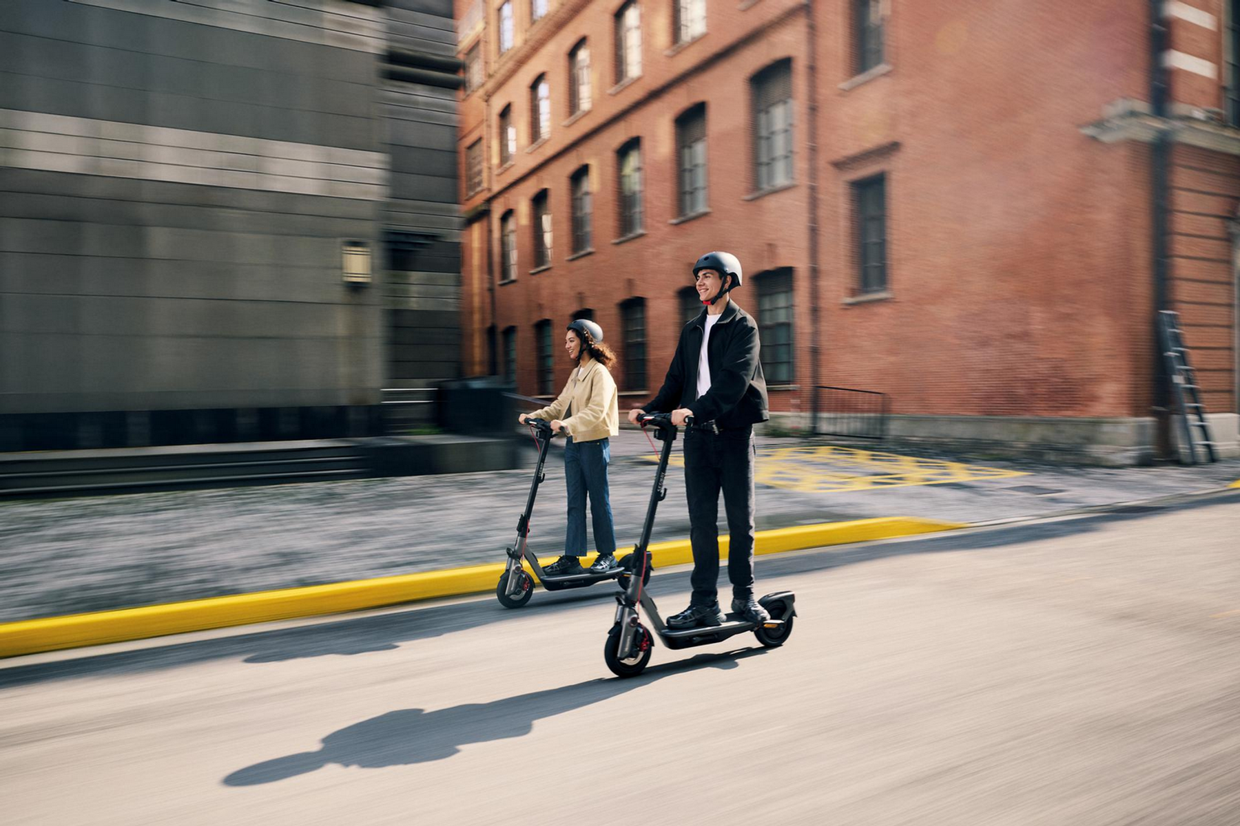 Zwei Personen fahren E-Scooter in einer Stadt mit Backstein- und modernen Gebäuden.