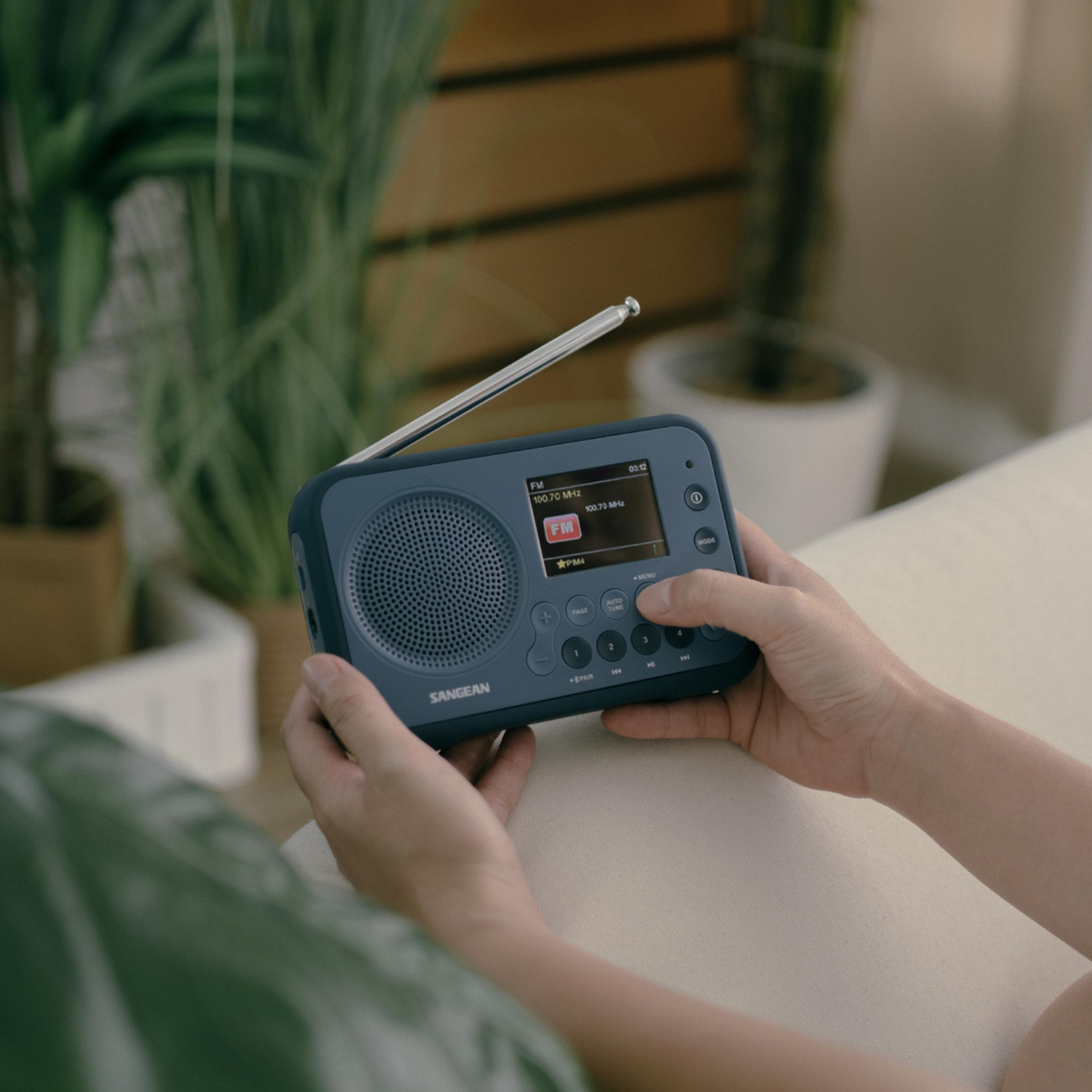 Une personne tient une radio Sangean bleue, l'écran affiche FM 100,70 MHz. Plantes en arrière-plan.