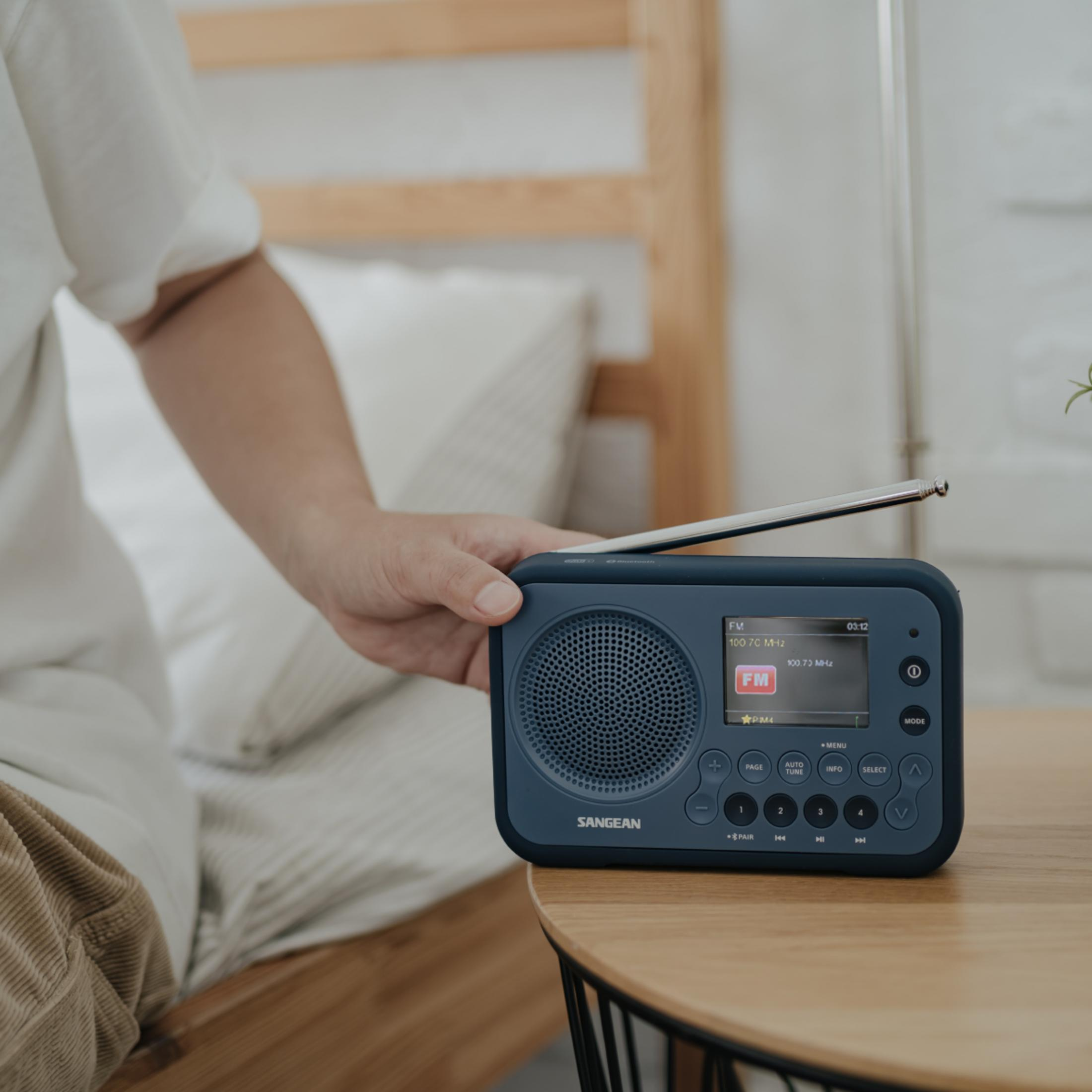Une main tient une radio Sangean bleue sur une table en bois à côté d'un lit.