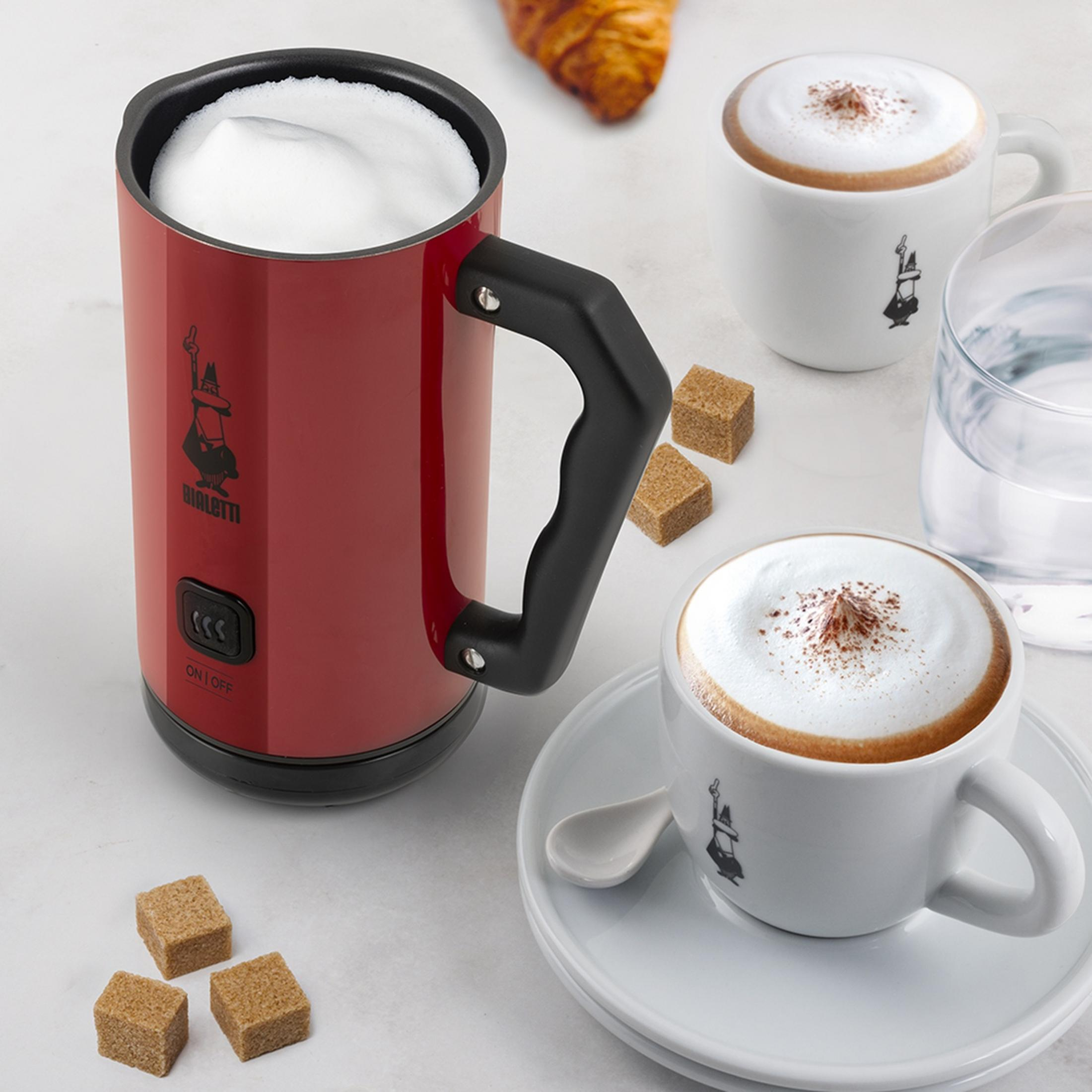 Czerwony spieniacz do mleka z pianką, dwa cappuccino, kostki cukru, szklanka wody i rogalik.