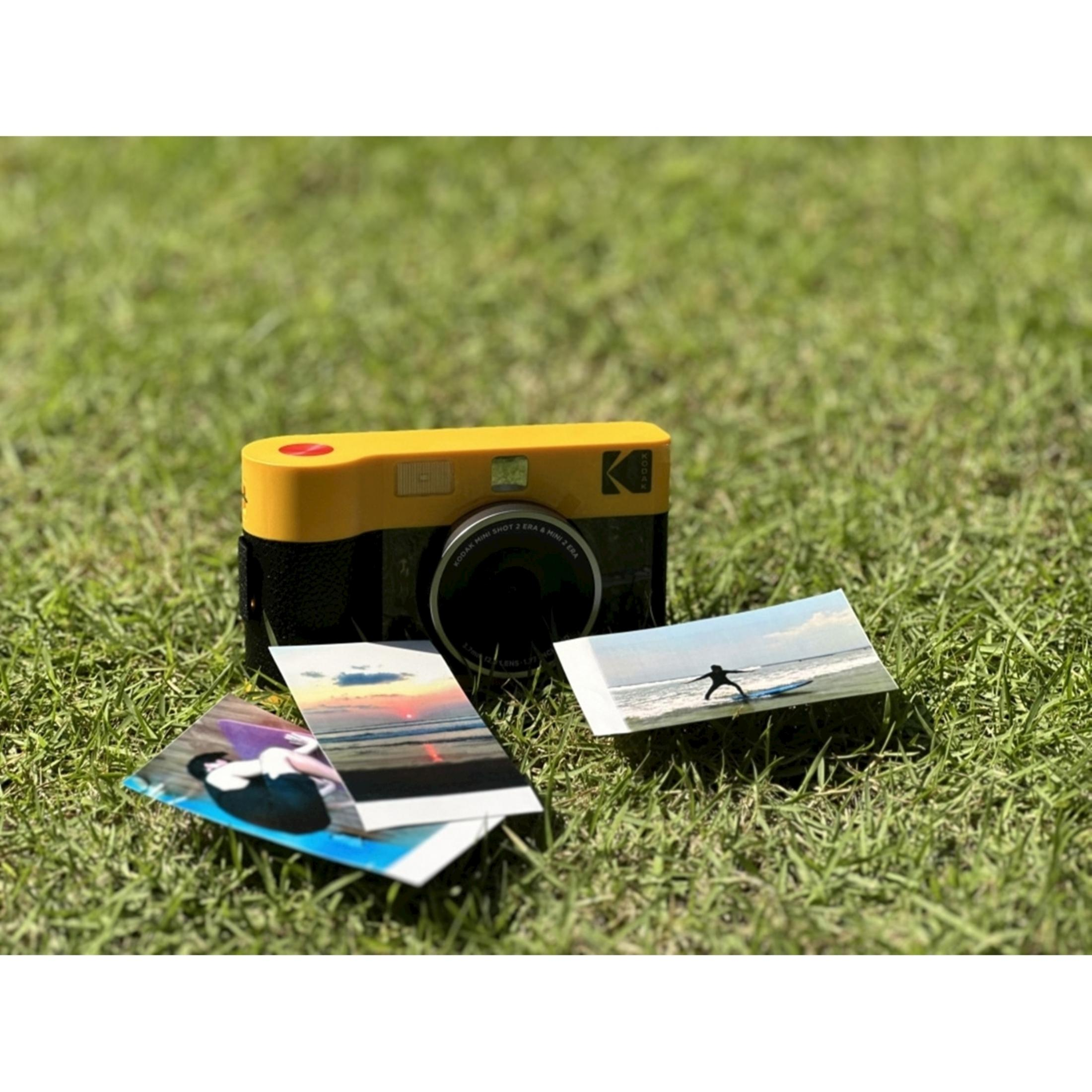 Ein gelber Kodak-Kameramini auf Gras mit drei Fotos davor.
