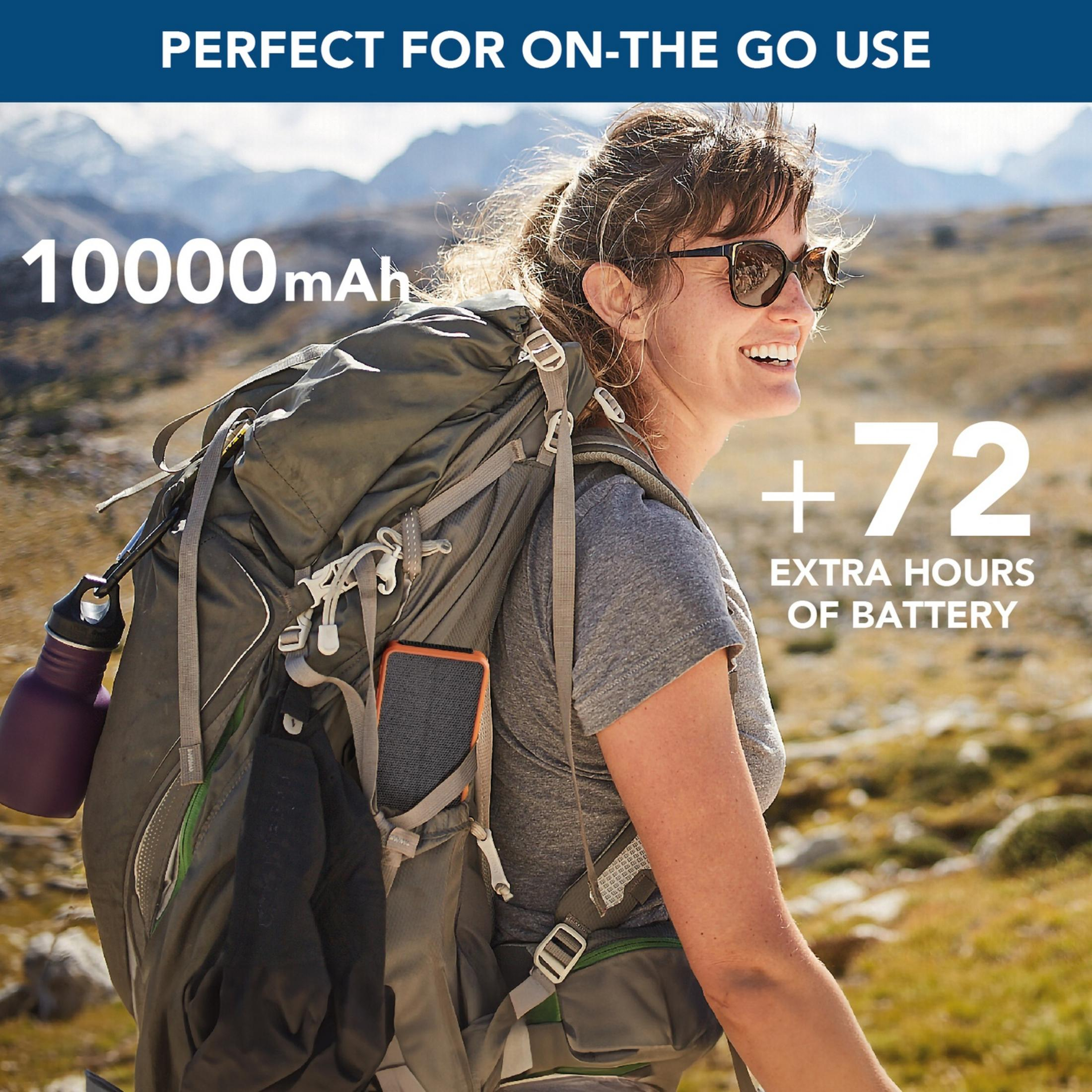 Une femme avec un sac à dos et des lunettes de soleil sourit, montagnes en arrière-plan.