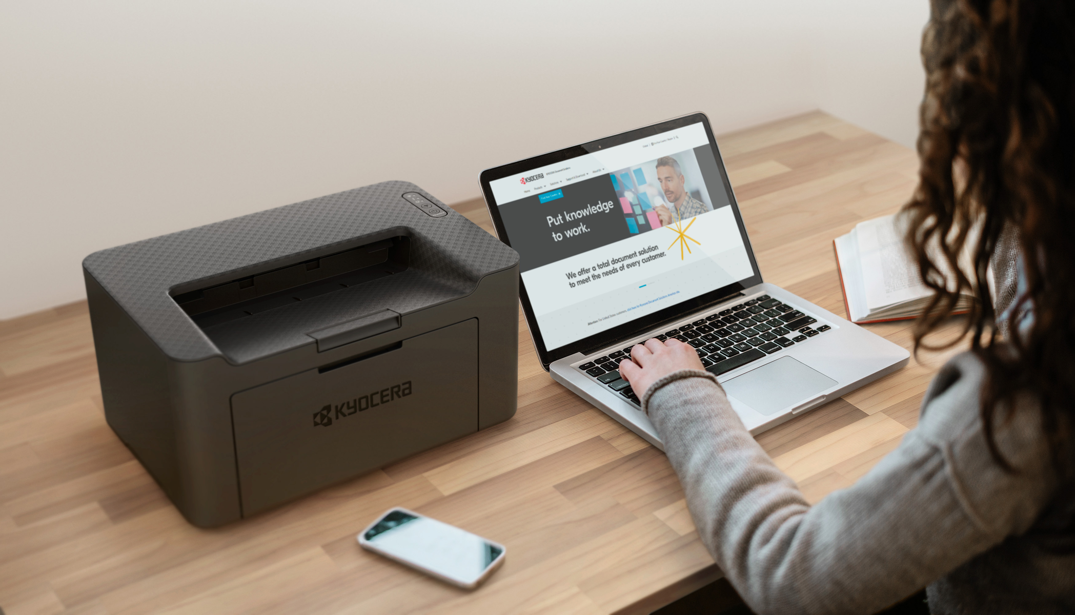 Una stampante e un laptop Kyocera su una scrivania di legno. Una persona digita sul laptop.