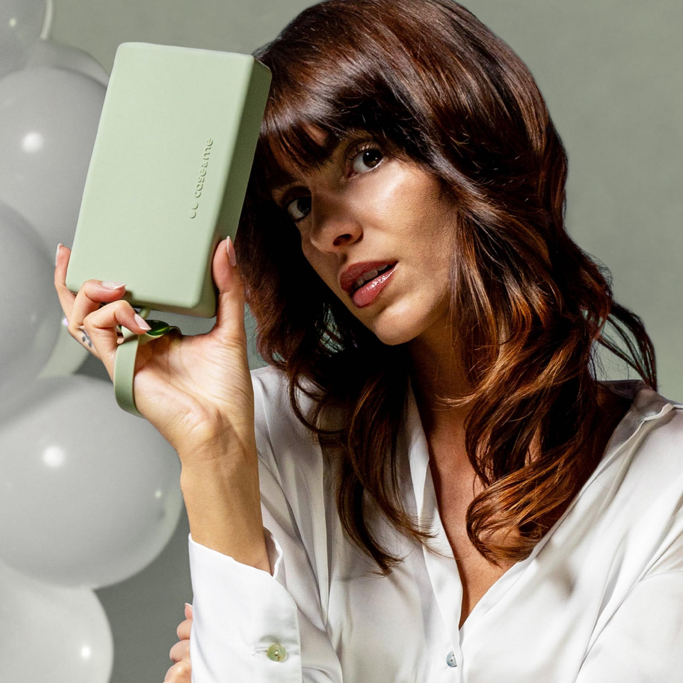 Donna con capelli castani tiene una custodia verde davanti al viso, palloncini bianchi sullo sfondo.