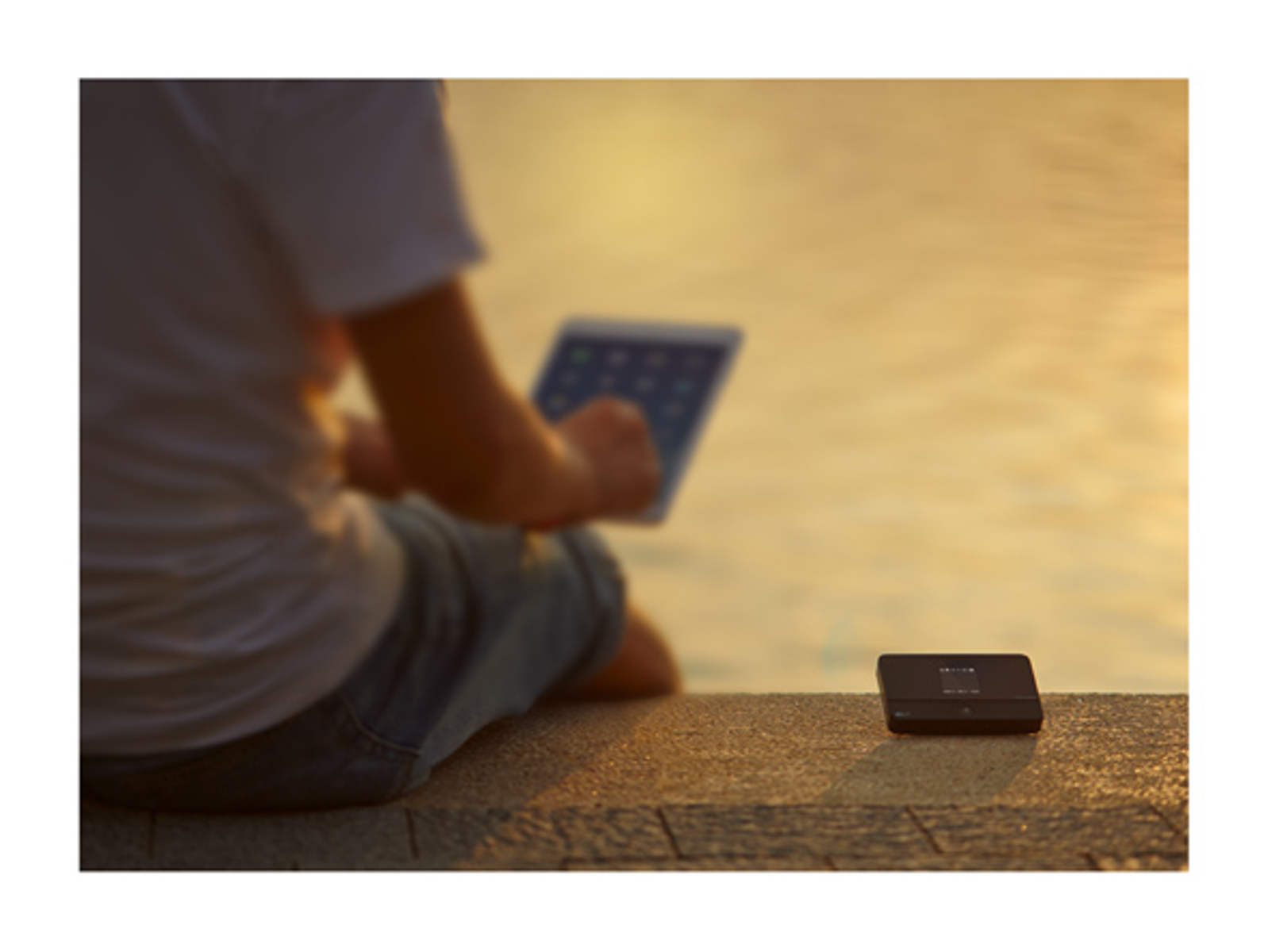 Eine Person sitzt im Freien und benutzt ein Tablet. Daneben liegt ein kleines schwarzes Gerät.