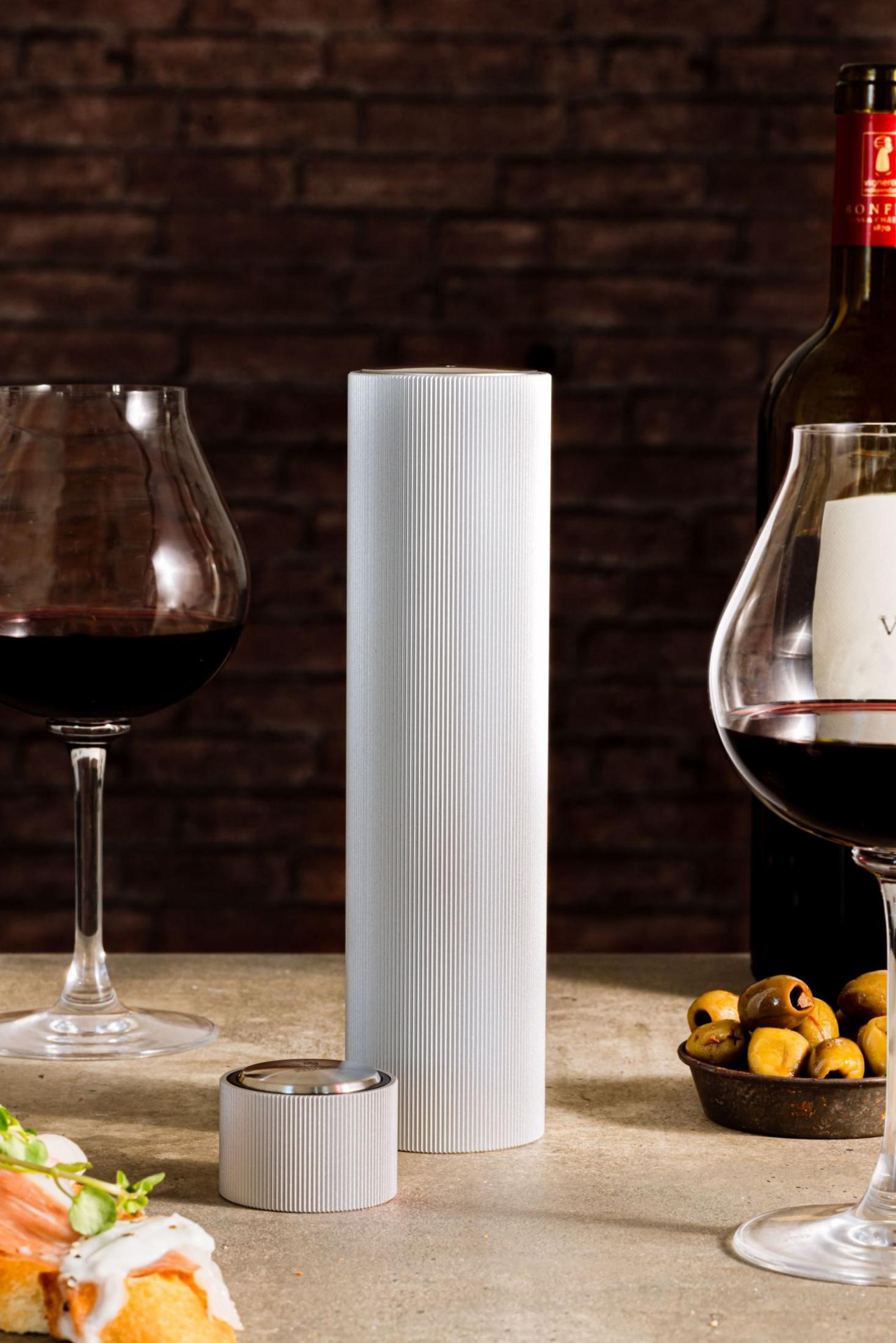 Ein silberner Zylinder steht zwischen zwei Weingläsern mit Rotwein. Oliven und Aperitifs sind zu sehen.