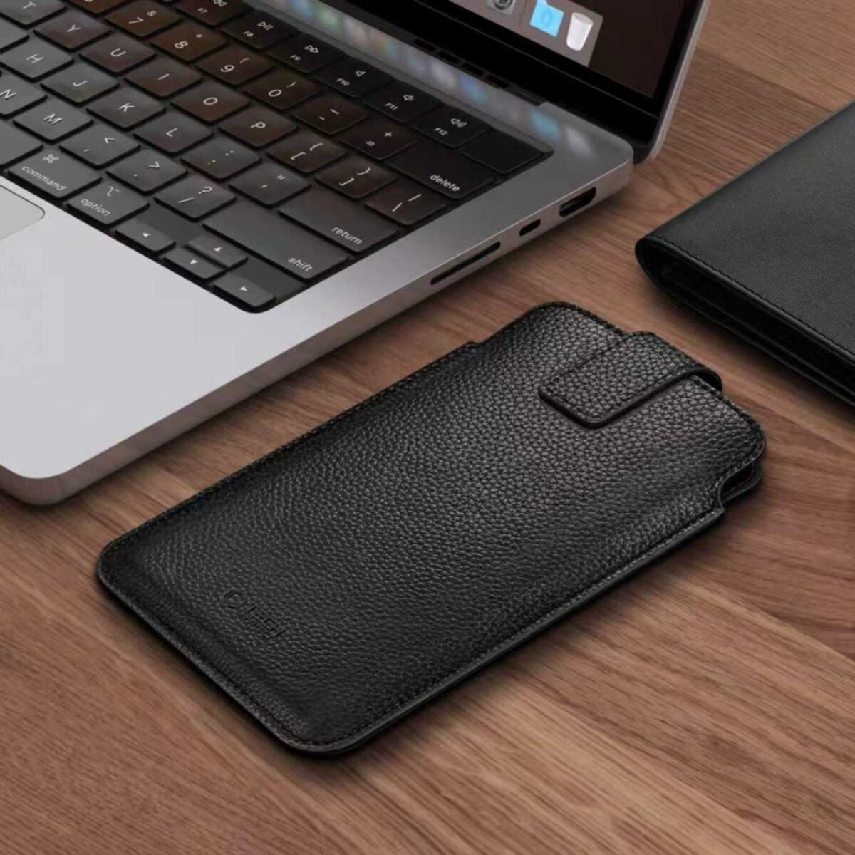 Eine schwarze Leder-Handyhülle liegt auf einem Holztisch neben einem Laptop.