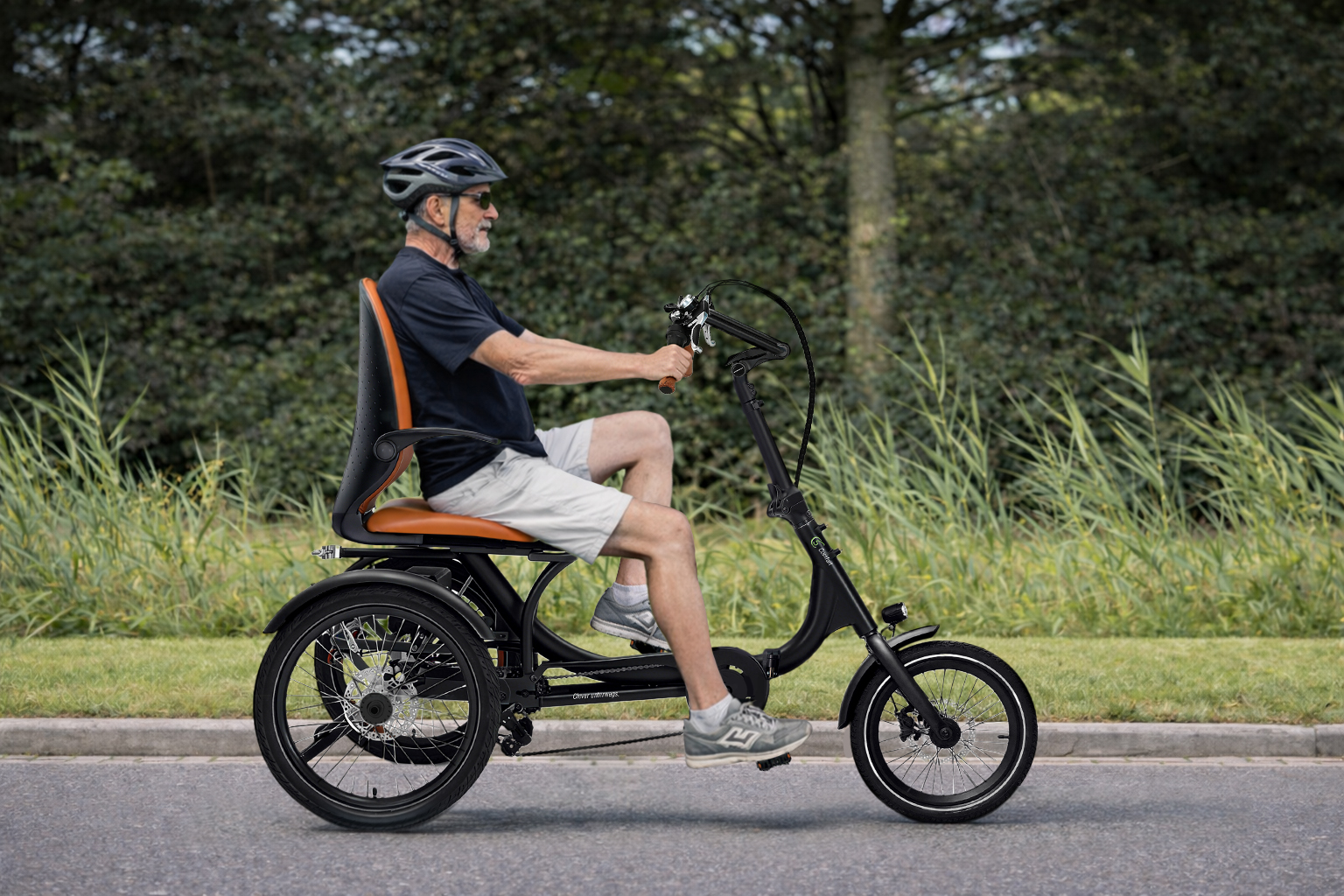 Ein älterer Mann mit Helm fährt ein schwarzes Dreirad auf einer Straße mit Bäumen.
