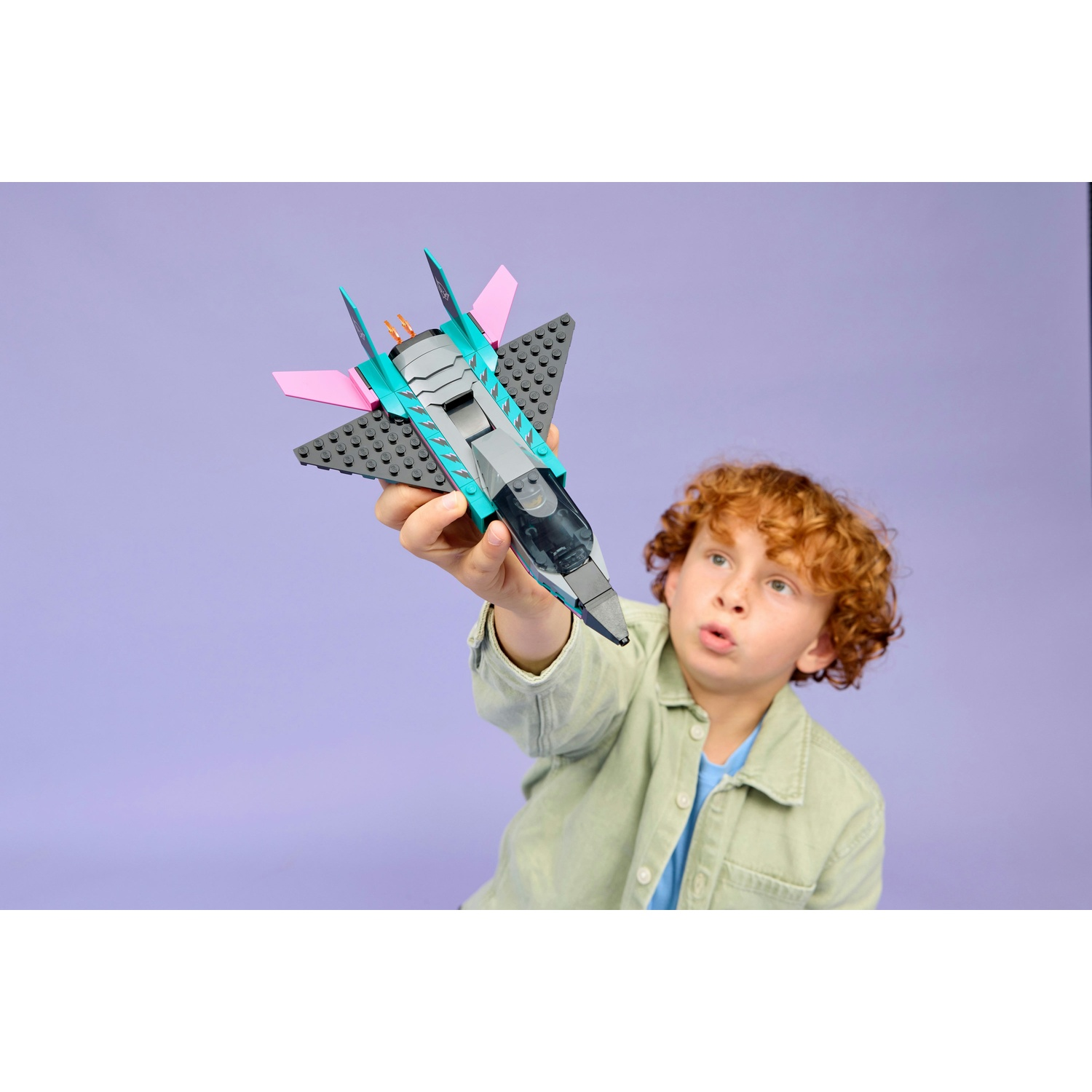 Chłopiec z rudymi włosami trzyma niebiesko-różowy samolot Lego na fioletowym tle.
