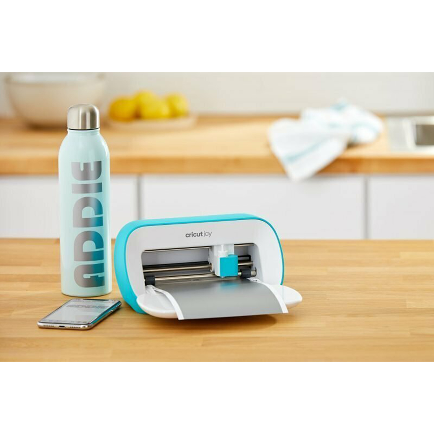 Ein blauer Wasserbehälter, ein Smartphone und ein Cricut Joy Schneidegerät auf einem Holztisch.