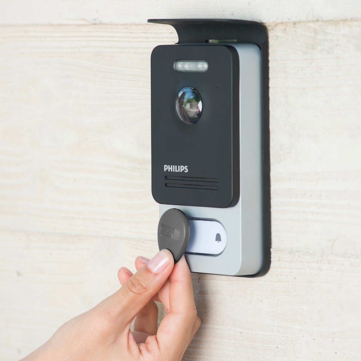 Una mano sostiene un llavero gris cerca de un timbre Philips con cámara.