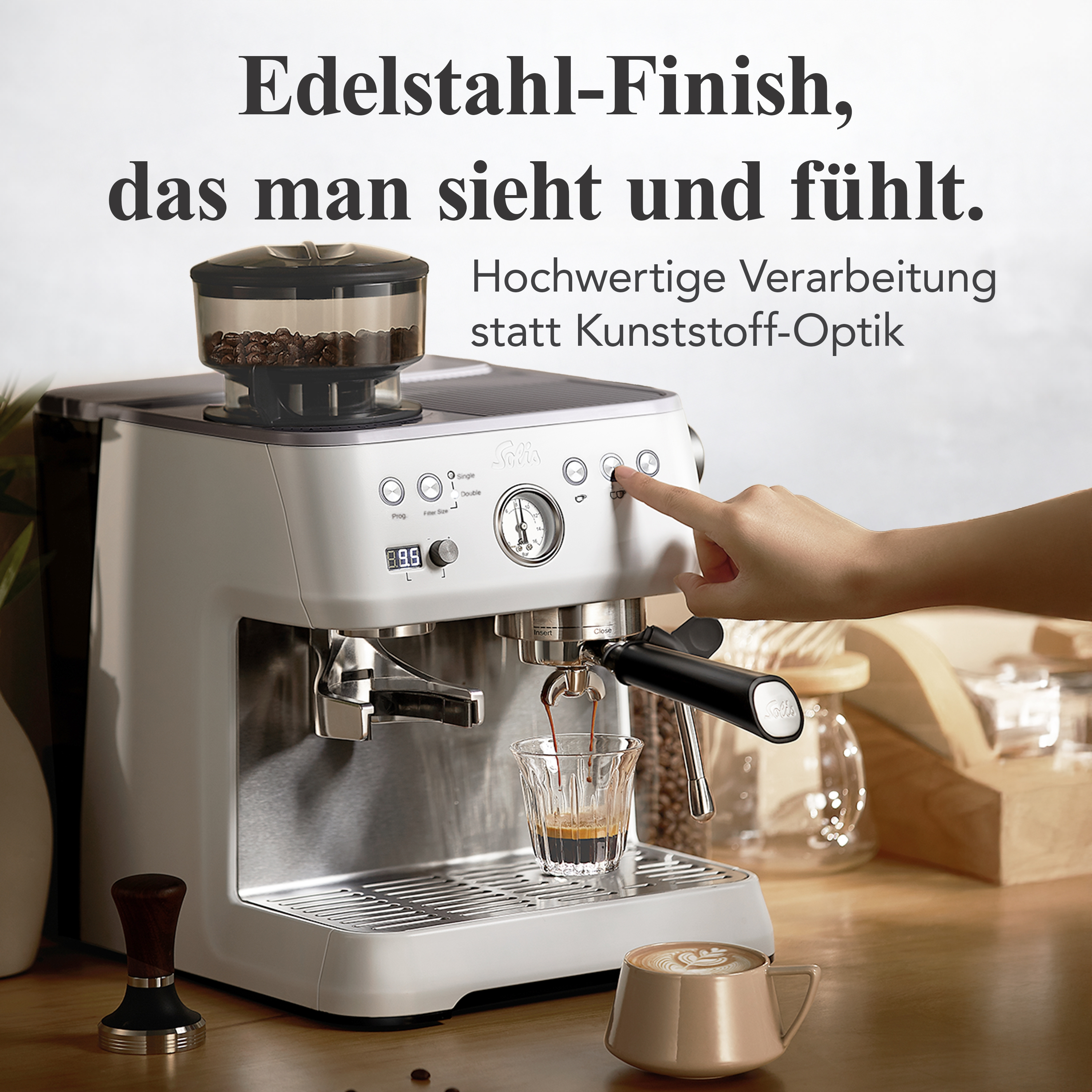 Eine weiße Espressomaschine brüht Kaffee in ein Glas. Ein Tamper liegt daneben.