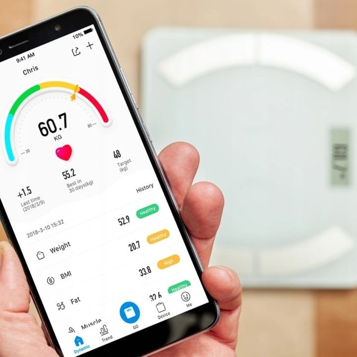 Una mano sostiene un smartphone con una app de salud que muestra el peso. Una báscula está al fondo.