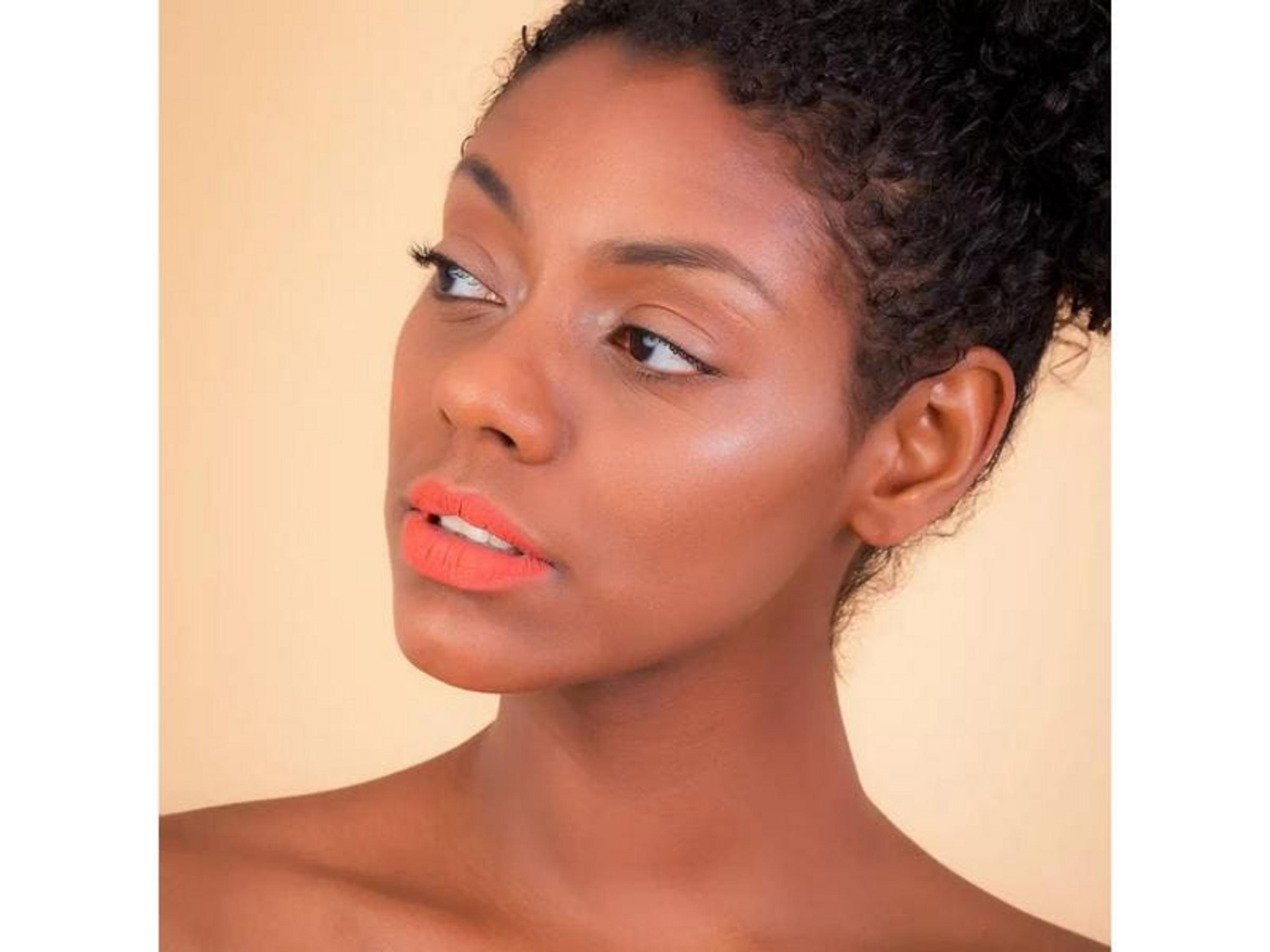 Nahaufnahme einer Frau mit orangefarbenem Lippenstift und lockigem Haar vor hellem Hintergrund.