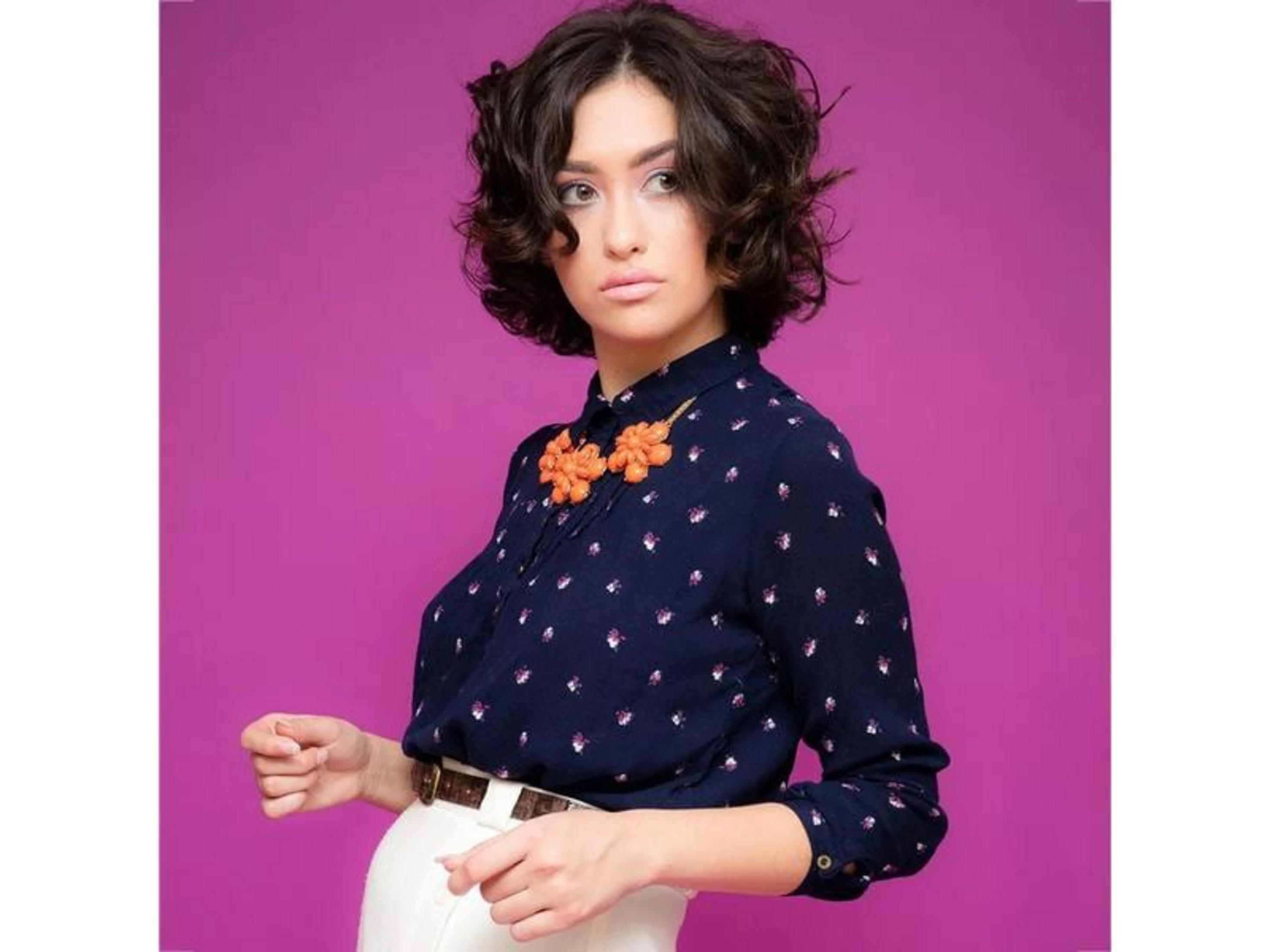 Une femme aux cheveux courts, foncés et bouclés porte un chemisier bleu marine à fleurs et un pantalon blanc.