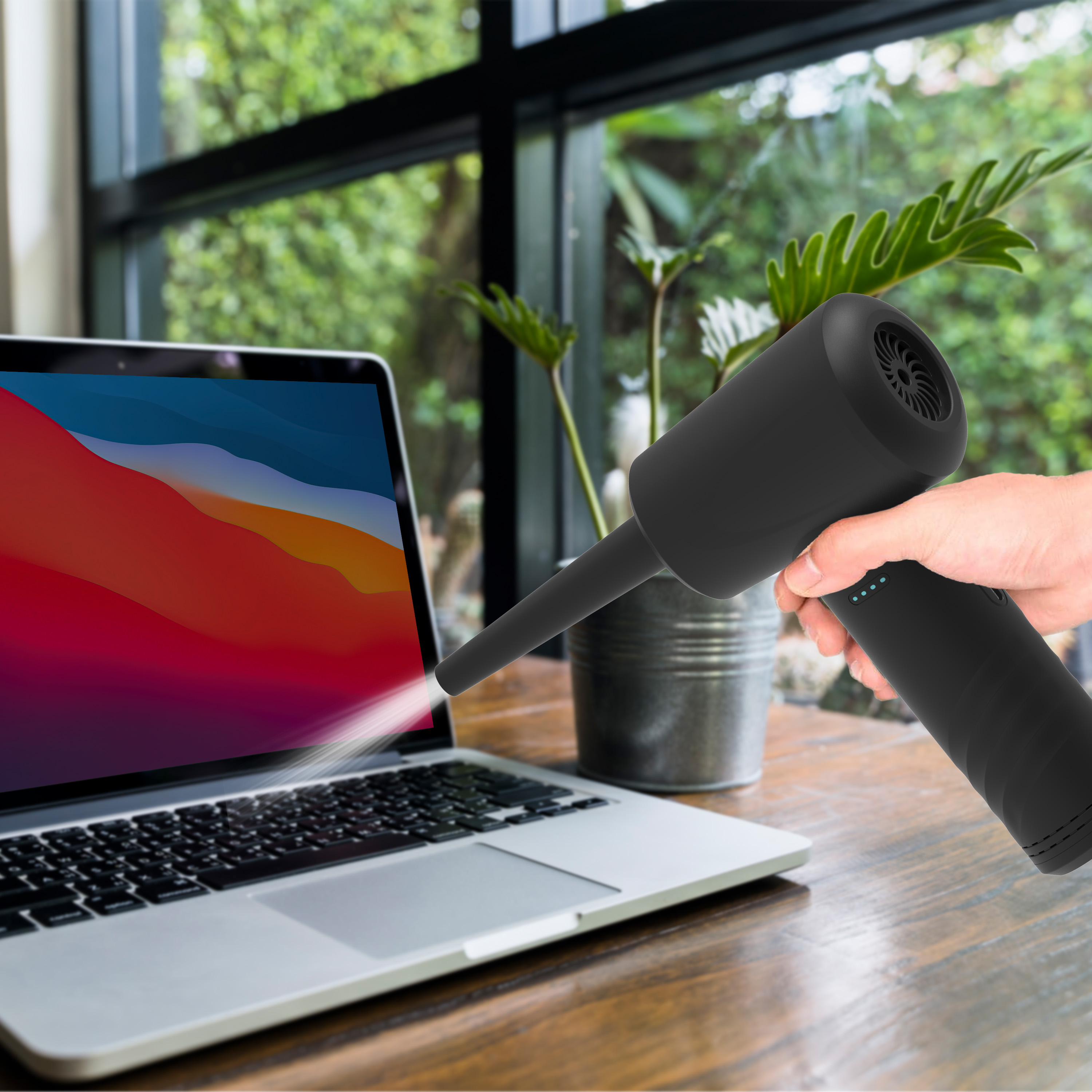 A hand holds a black blower aimed at a laptop. Outdoors in background.